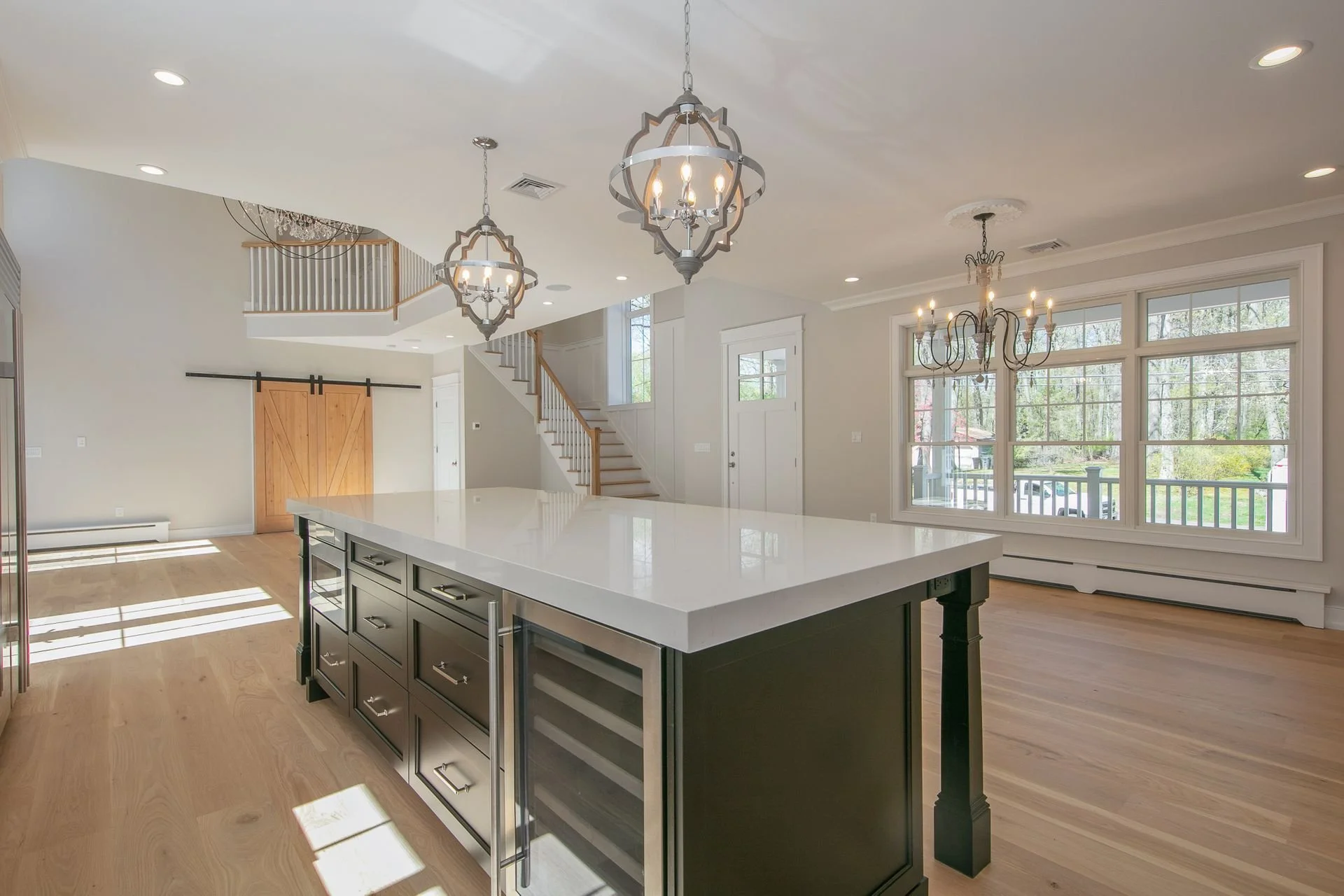 Bright open kitchen with large island, black cabinetry, modern chandeliers, and large windows overlooking a green outdoor space.