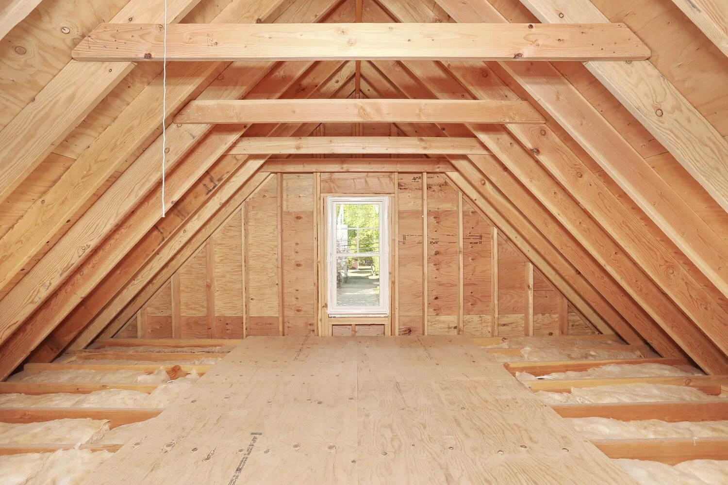 Interior of an unfinished attic with exposed wooden framing, insulation, and a small window at the end.