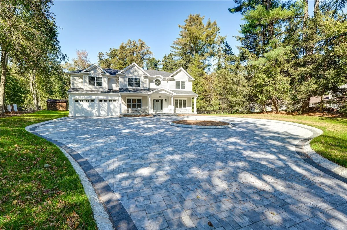 Large white two-story house with a detached garage, surrounded by green trees and a landscaped front yard with a paved driveway and a circular planting bed in the center.