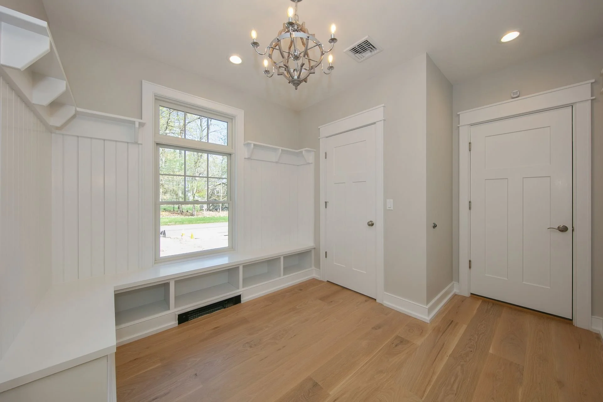 Interior of a bright room with white walls, a large window, wooden flooring, a chandelier hanging from the ceiling, and two white doors with trim.