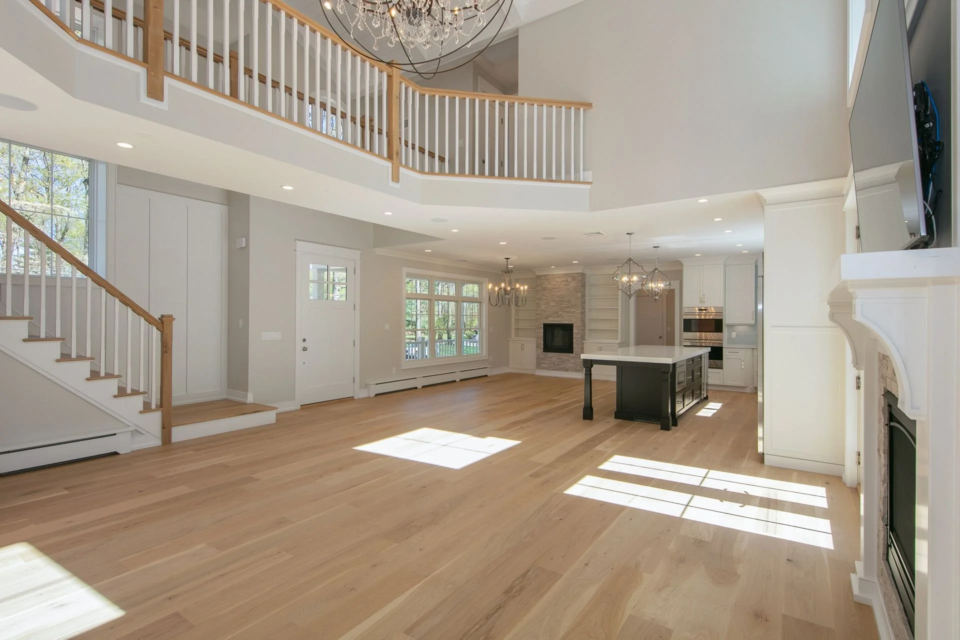 Spacious living room with hardwood floors, large windows, a fireplace with stone surround, white cabinetry, a kitchen island, and modern chandeliers. Upstairs balcony with wooden railing overlooks the area.