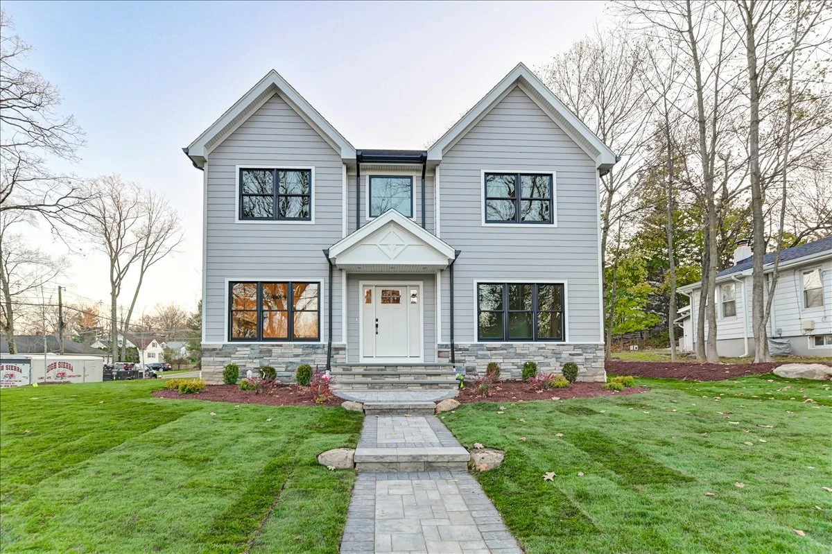 Front view of a two-story house with gray siding, white trim, and a small front porch with steps leading to the front door. The house has three large windows on each floor and is surrounded by a well-maintained green lawn and trees.