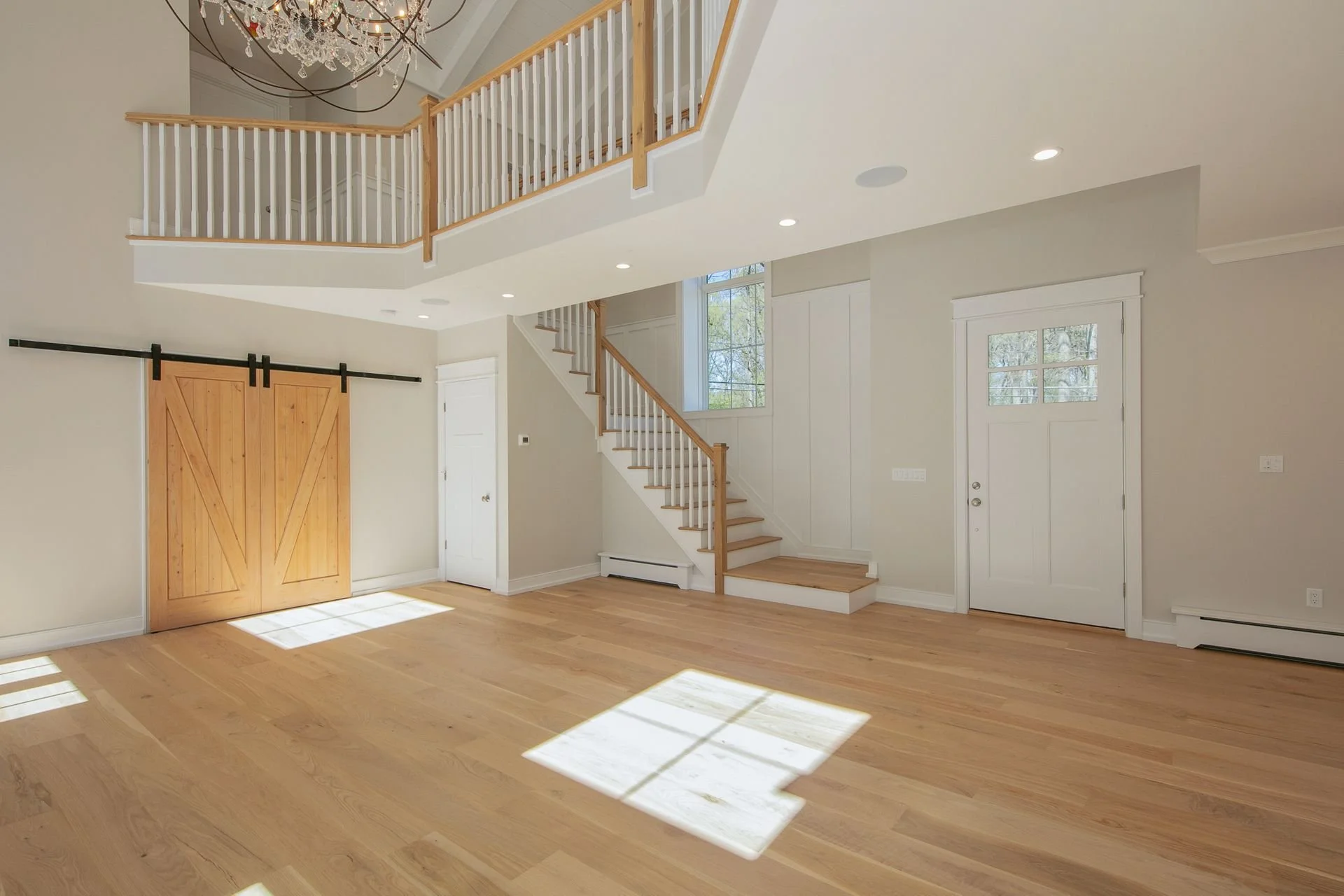 Interior of a modern home with hardwood floors, white walls, staircase with wooden railing, sliding barn door, large windows, and chandelier hanging from high ceiling.