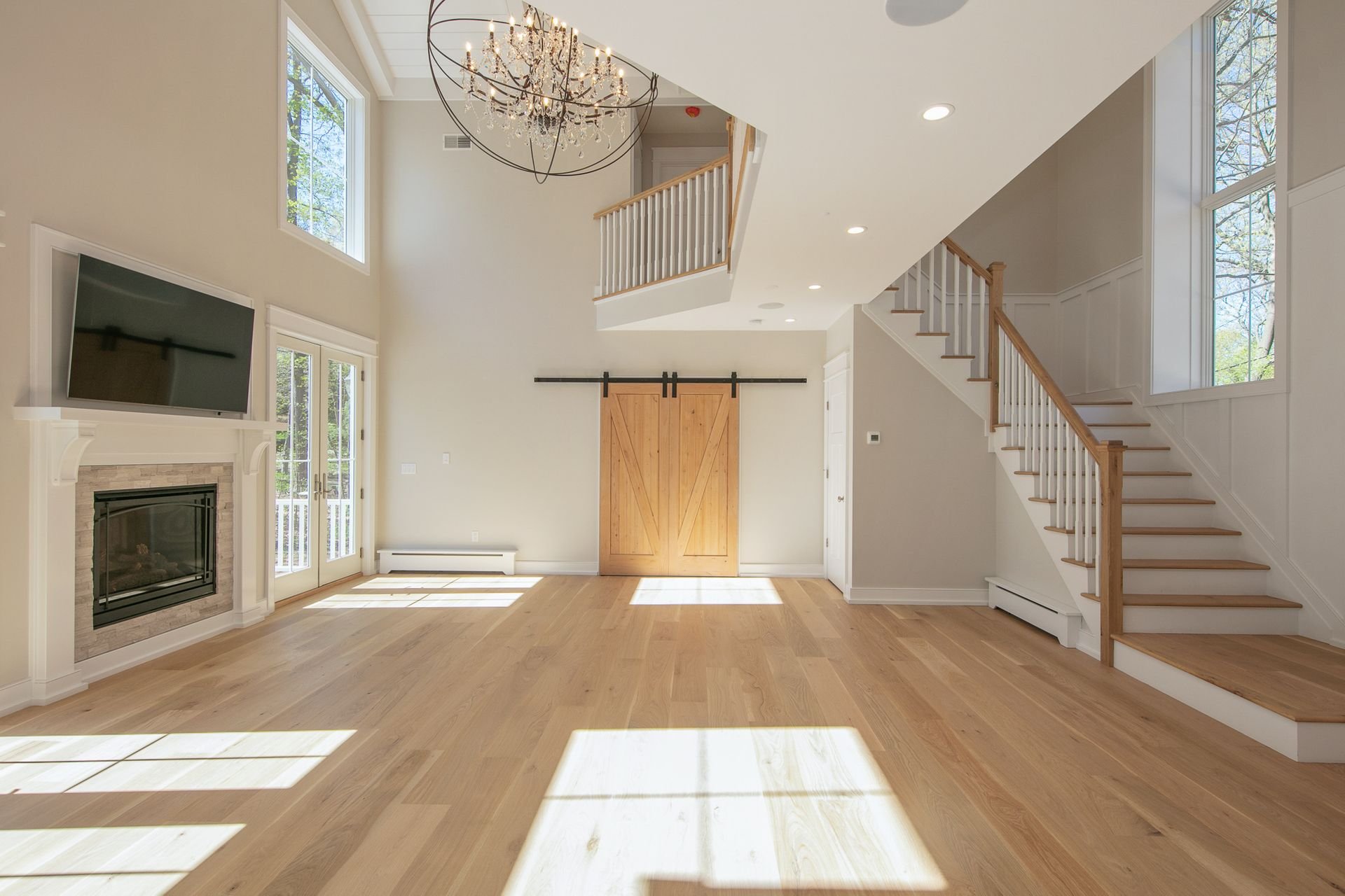 Bright living room with large windows, a chandelier, a fireplace with a mounted TV above, a sliding glass door, a wooden barn door, and a staircase with white railing and wooden steps.