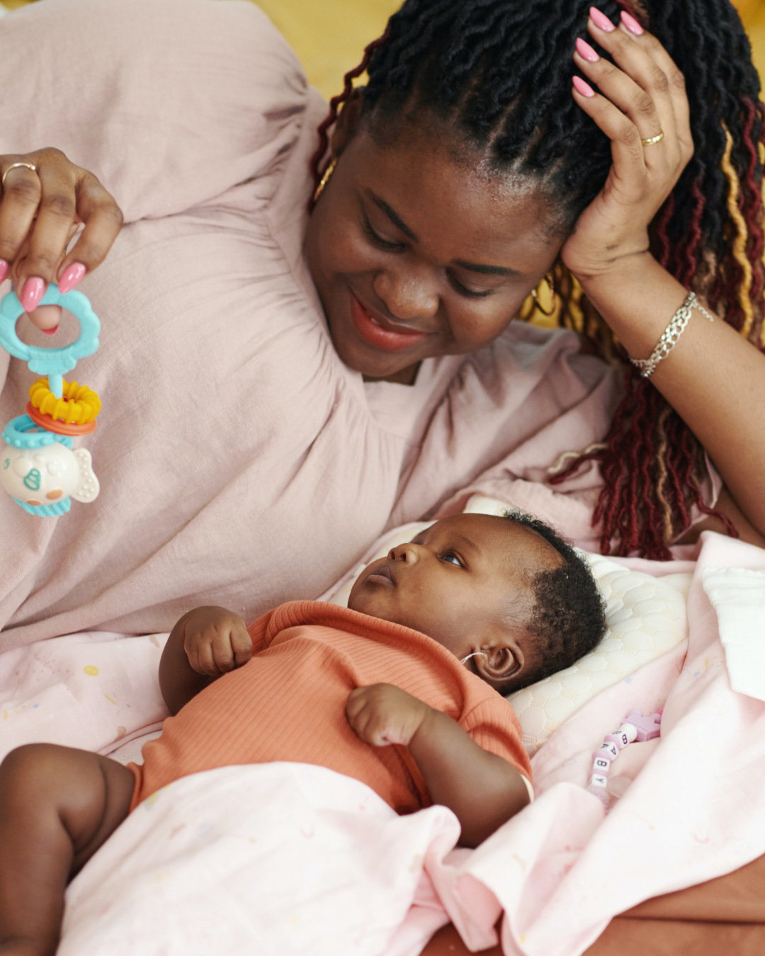 A woman with colorful braids and pink nails looks at a baby lying on a bed, holding a pacifier toy.