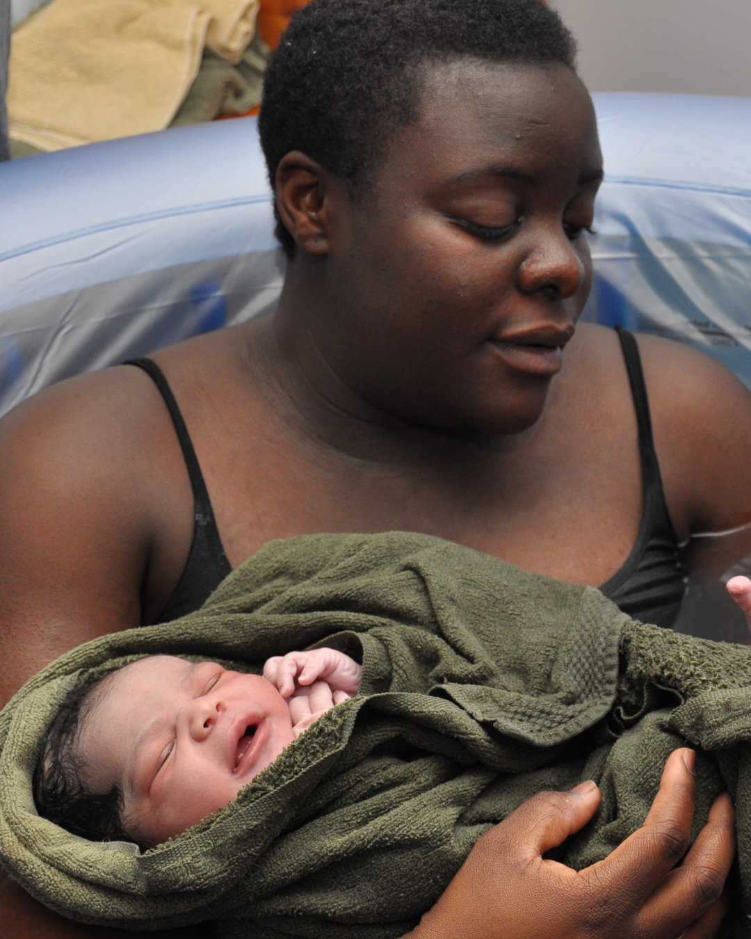 A woman holding a newborn baby wrapped in a green towel in a birth pool in her home..