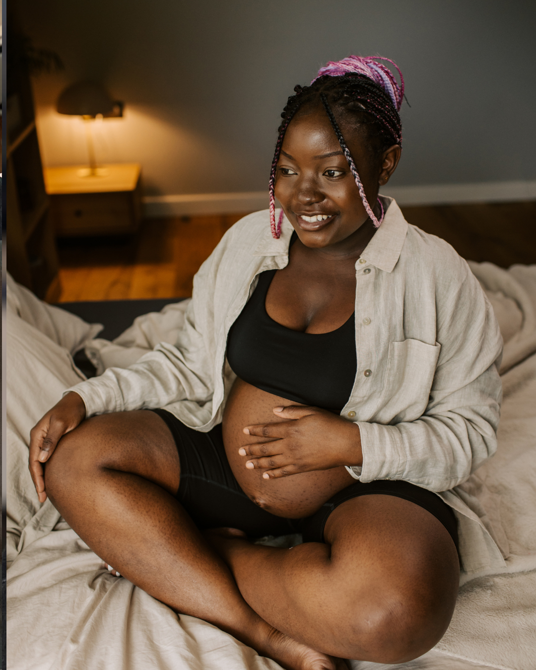 A pregnant woman sitting cross-legged on a bed, smiling, touching her belly. She has dark skin, braided hair with pink highlights, and is wearing a black top, beige shirt, and black shorts. There is a nightstand with a lamp in the background.