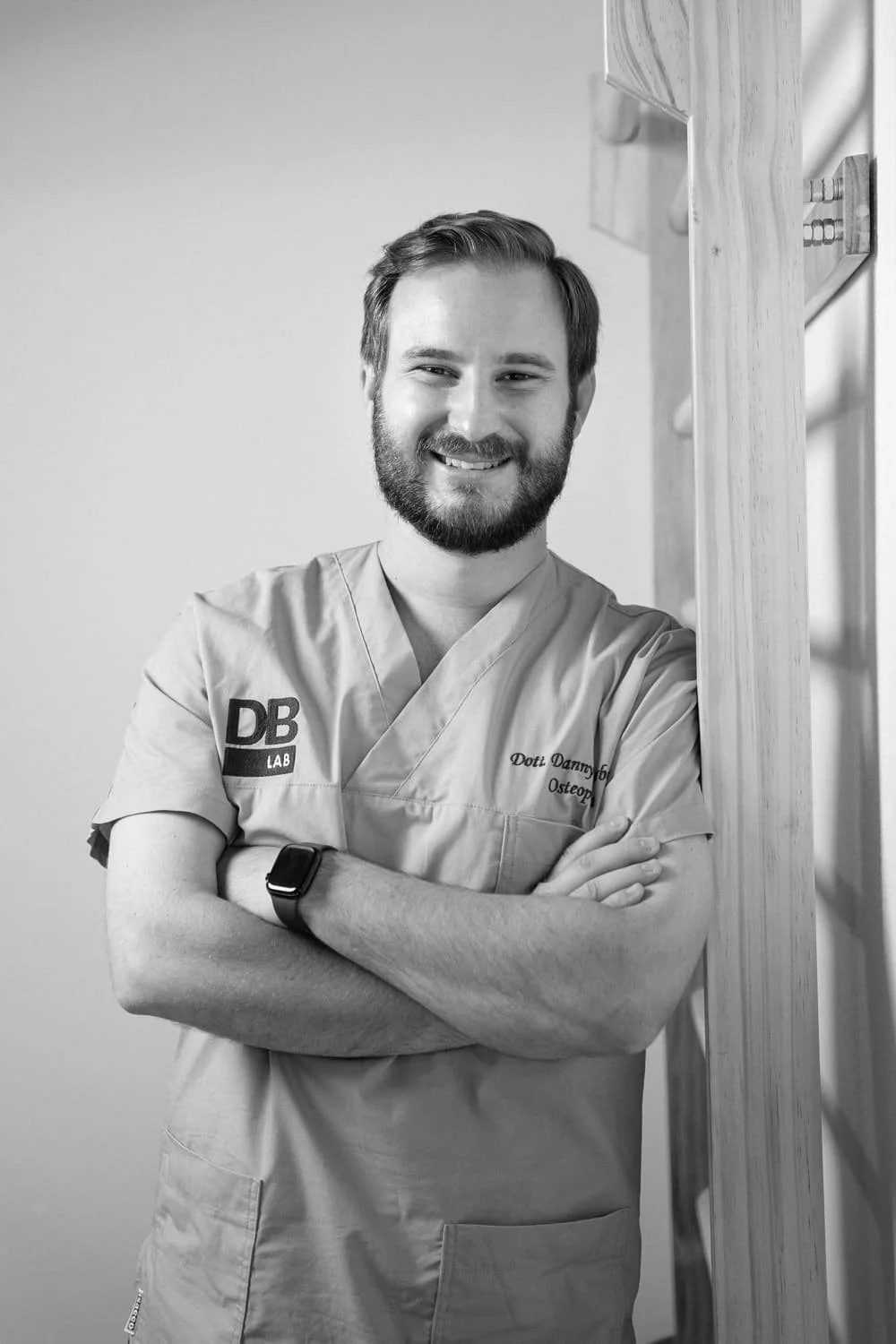 Black and white photo of a smiling male healthcare professional standing with arms crossed in front of a wooden structure. He is wearing scrubs with the embroidered name Doti Danny Boy, Osteo, and a watch on his left wrist.