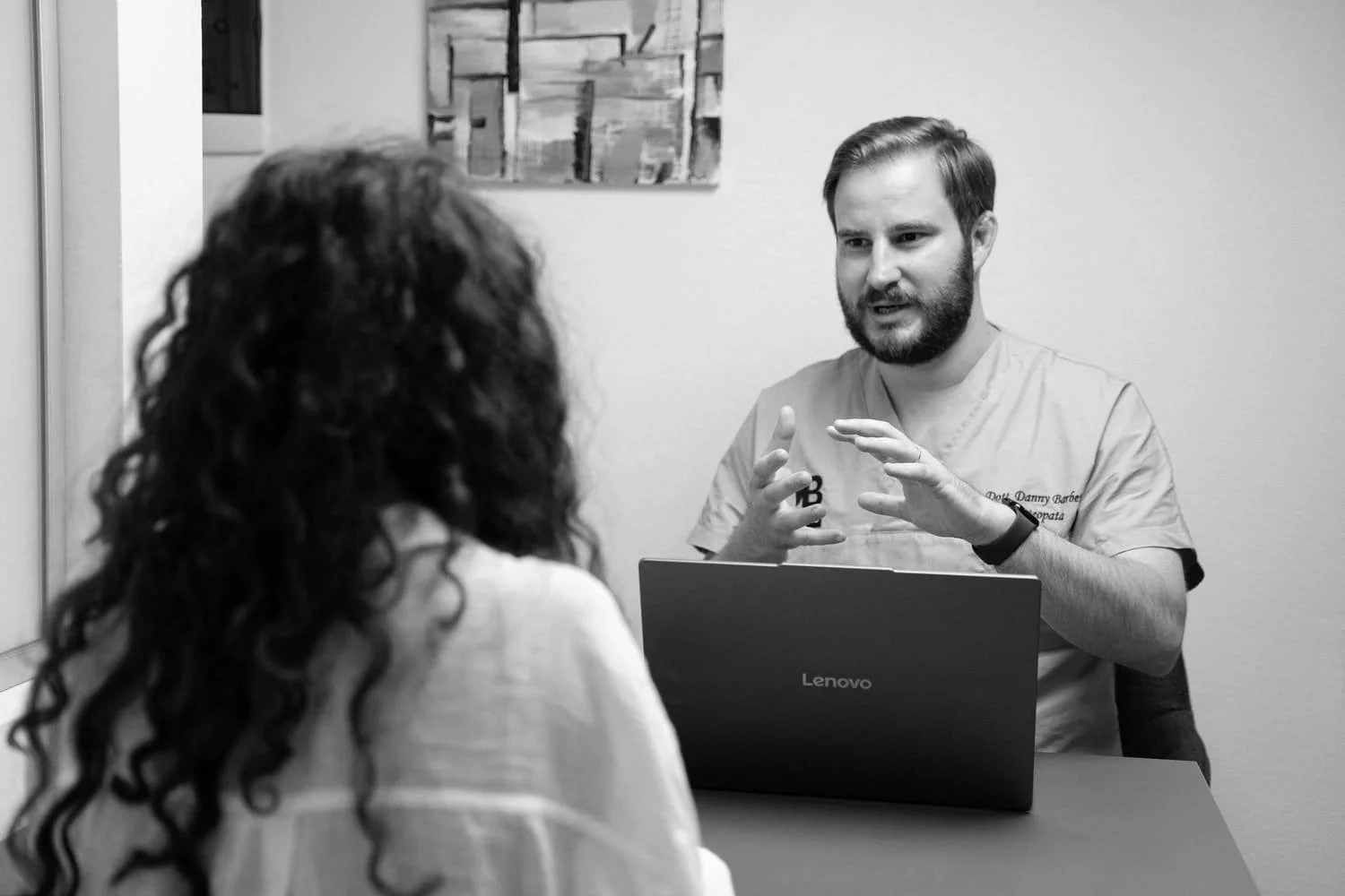 A man and a woman in a medical office or consultation room. The man, wearing scrubs, is sitting behind a desk with a Lenovo laptop open in front of him. He is gesturing with his hands while speaking to the woman, who has curly hair and is sitting across from him.