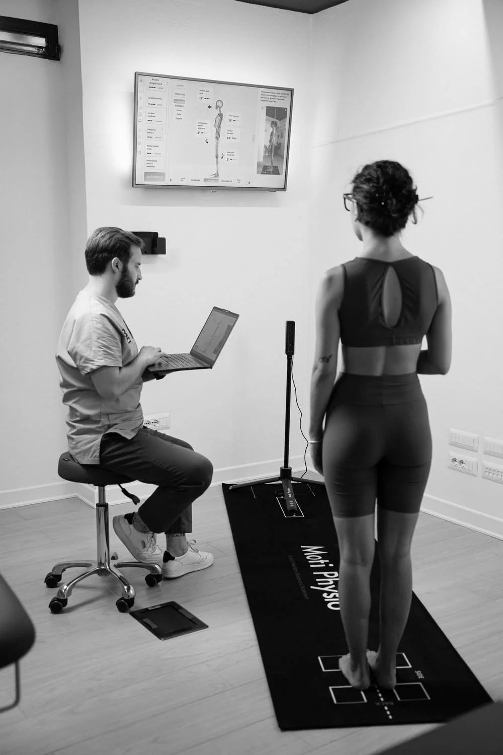 A woman standing on a motion analysis mat in a biomechanics lab, with a technician sitting on a stool using a laptop, while a monitor displays a digital image of a human body with measurements