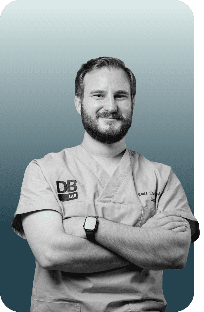 A smiling man with a beard and short hair in medical scrubs, crossing his arms, against a gradient background.