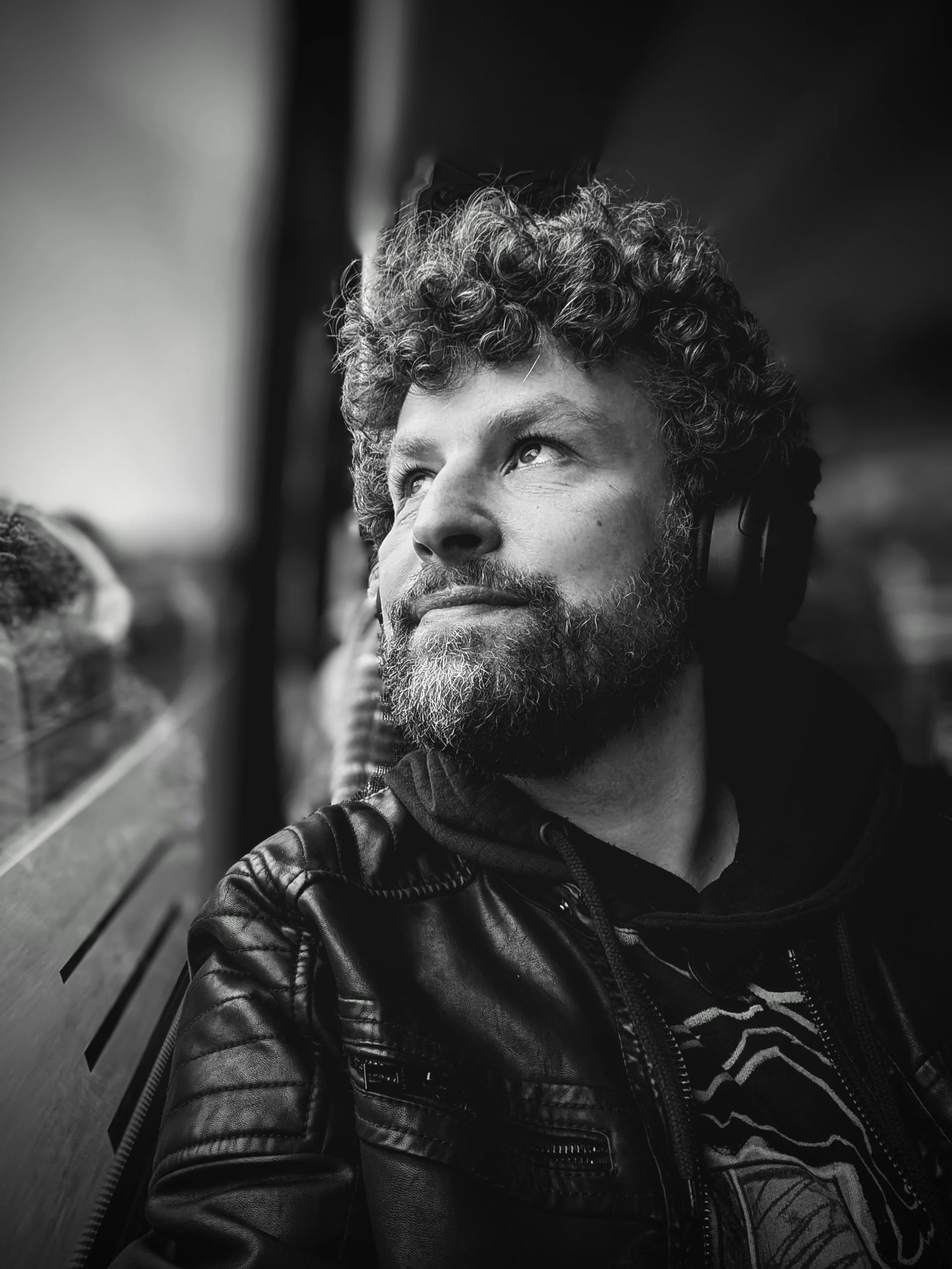 Black and white photo of a man with curly hair and a beard, looking up while sitting on a wooden bench. He is wearing a leather jacket and a hoodie with a design visible on it.