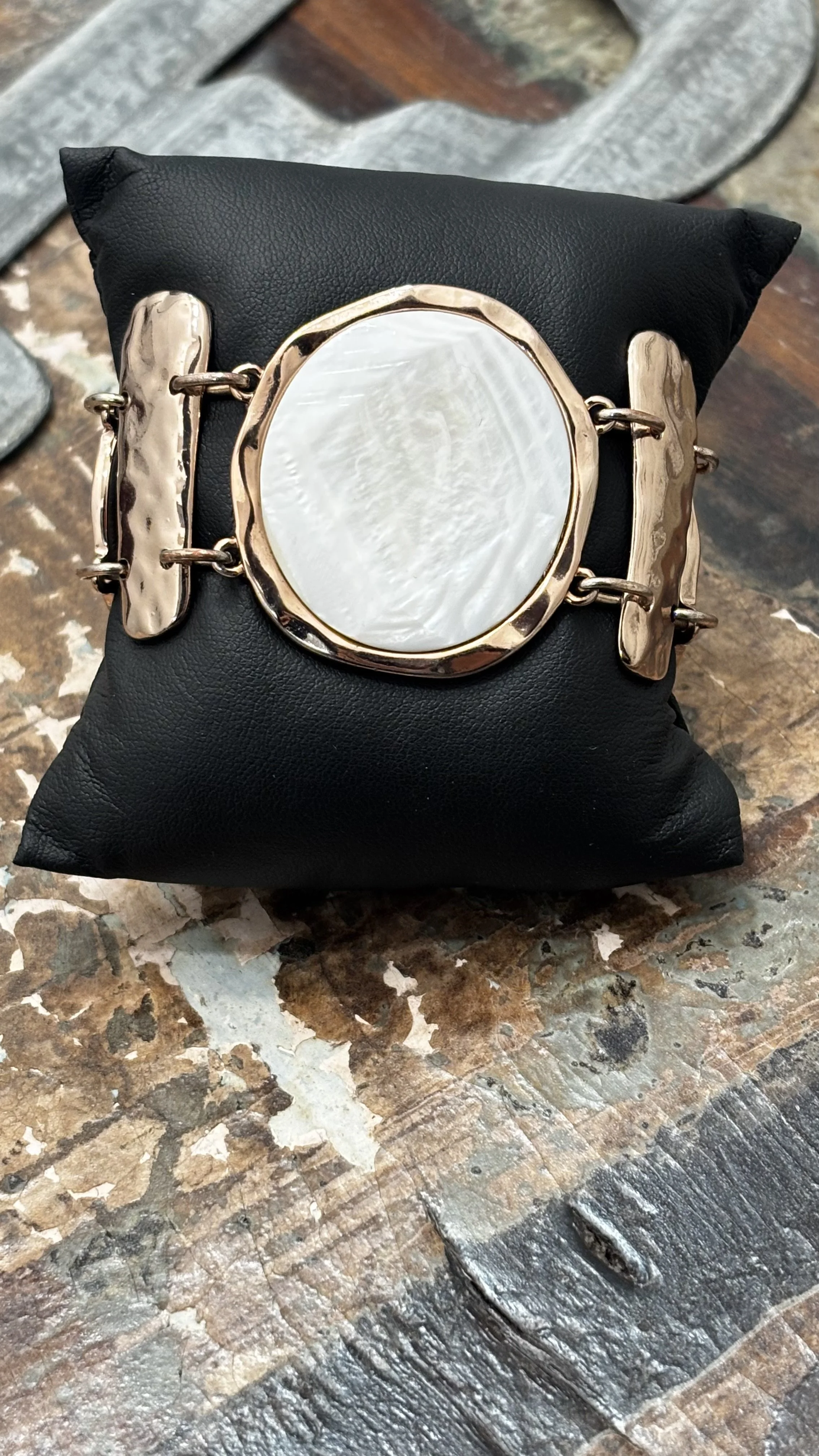 A black cushion with a decorative bracelet featuring a large oval white shell set in a gold-tone frame, placed on a rustic wooden surface.