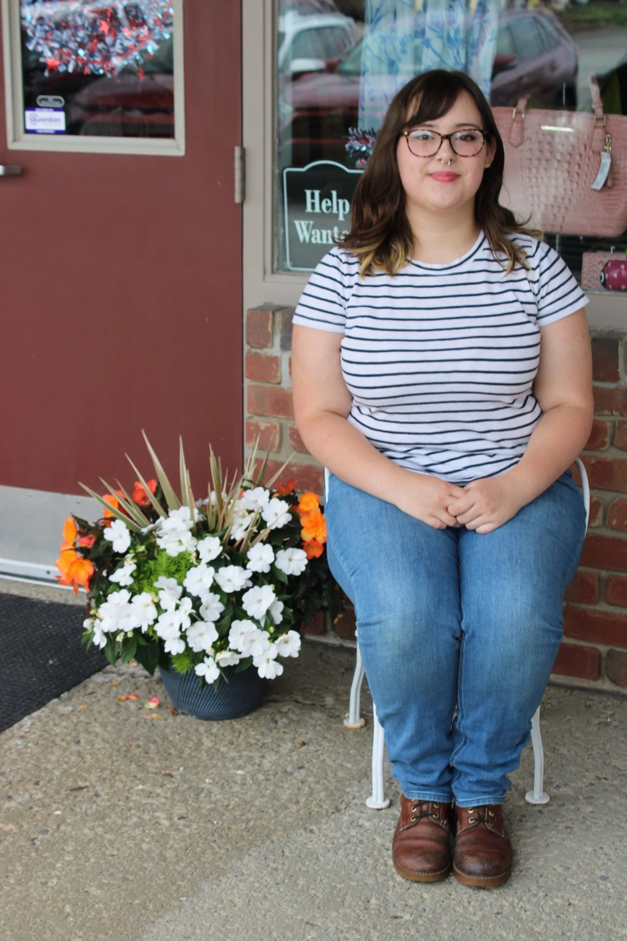 A young woman with glasses and a septum piercing sitting on a white chair outside a store, smiling. She is wearing a striped t-shirt and blue jeans. Next to her, there is a flower planter with white, orange, and green flowers. The store window behind her displays pink handbags and a sign that reads 'Help Wanted.'