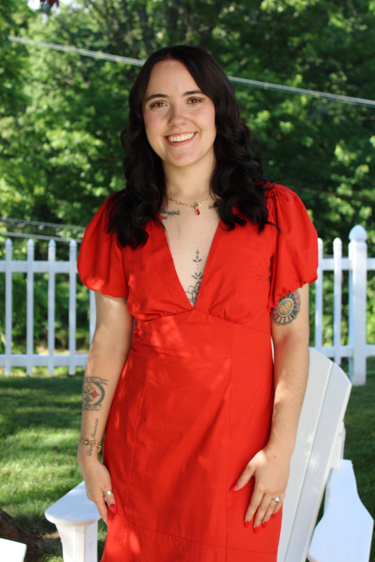 A woman with dark wavy hair wearing a red dress and jewelry, standing outdoors in front of a white fence and lush green trees.