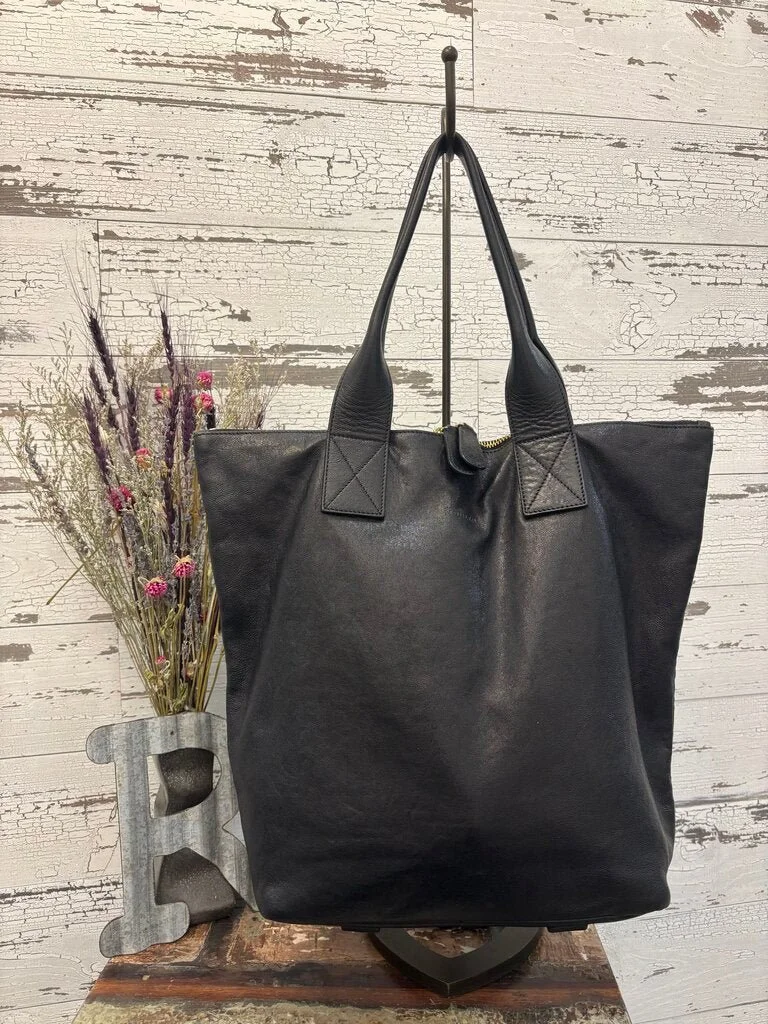 Black leather tote bag hanging on a hook, placed on a rustic wooden surface, with a decorative letter 'E' and a dried floral arrangement nearby against a distressed white wooden background.