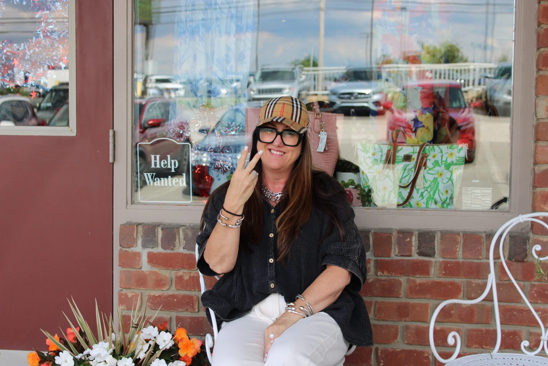A woman with long brown hair, black glasses, wearing a black shirt and white pants, sitting outside in front of a brick wall and a window of a store, smiling and making a peace sign with her fingers.