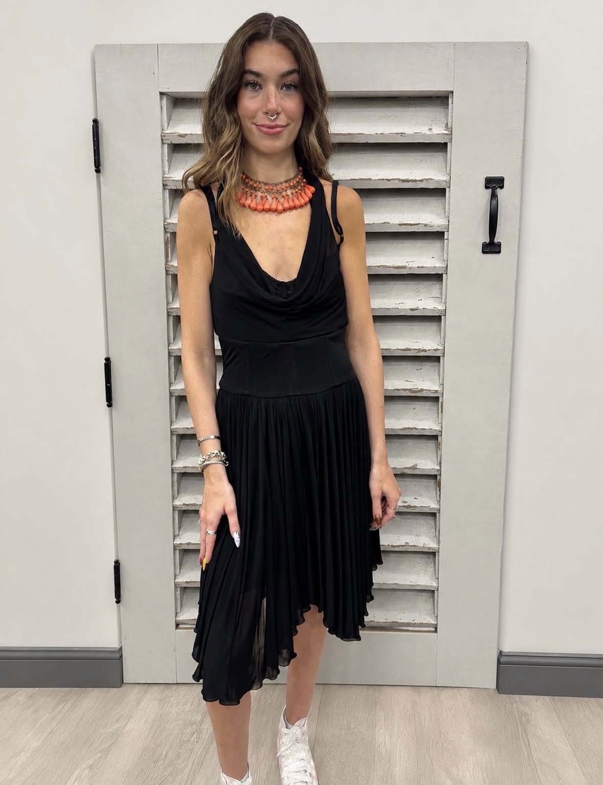 A young woman with wavy brown hair, wearing a black dress with a pleated skirt, silver jewelry, orange necklace, white sneakers, standing in front of a white wooden door with horizontal slats.