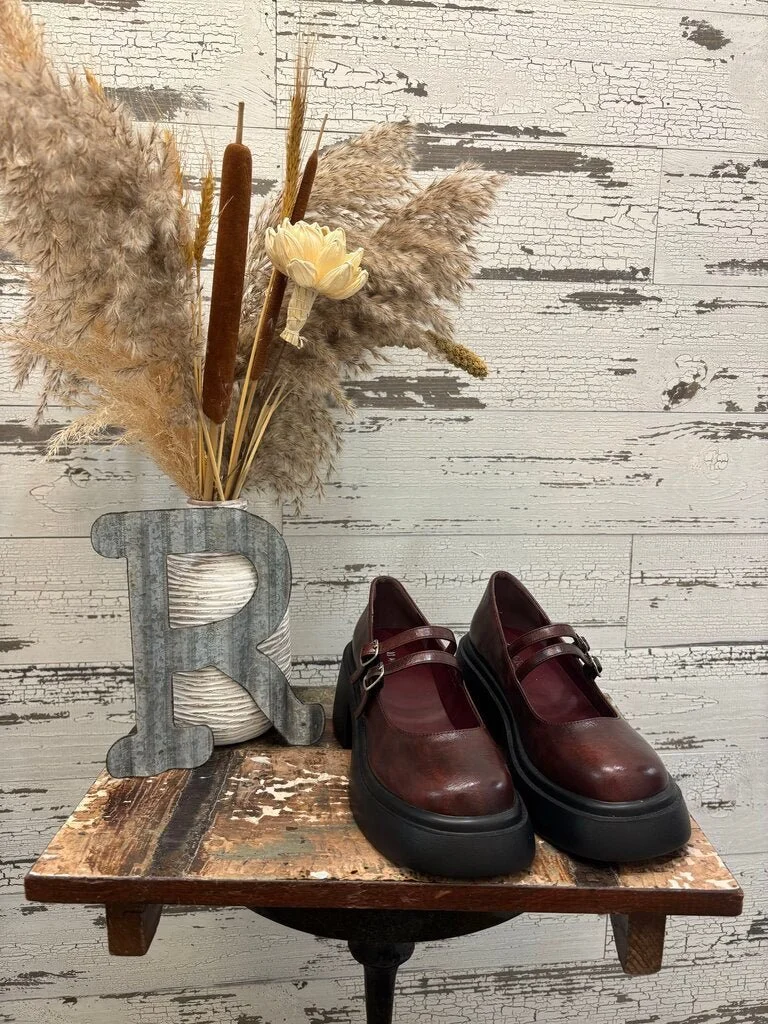 Pair of burgundy leather platform shoes with ankle straps on a rustic wooden table, with a decorative metal letter 'R' and a vase with dried pampas grass and flowers in the background.
