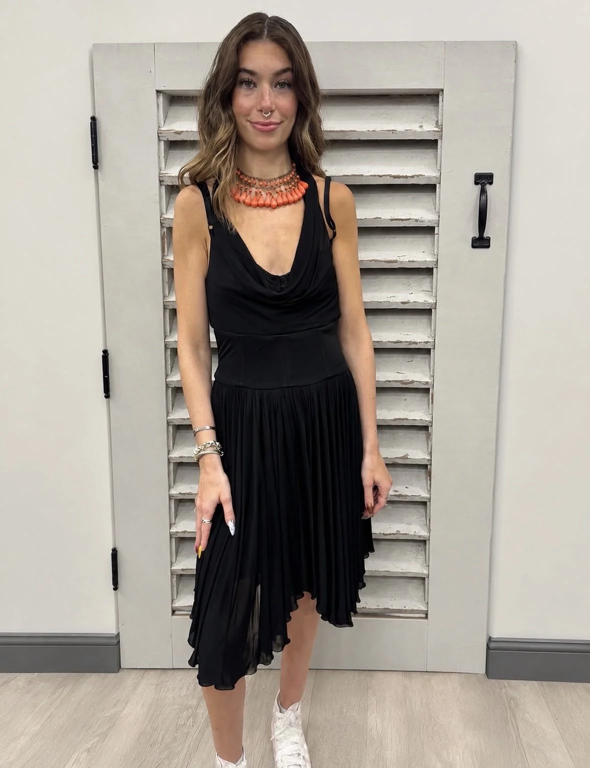 A young woman with wavy brown hair wearing a black dress, white sneakers, and colorful jewelry, standing in front of a white wooden wall.