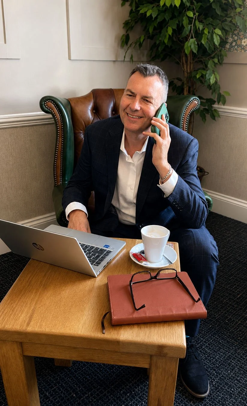 Andy sitting at a wooden table, talking on a cellphone to a customer, with a laptop, a white coffee cup with a saucer, and a notebook on the table.