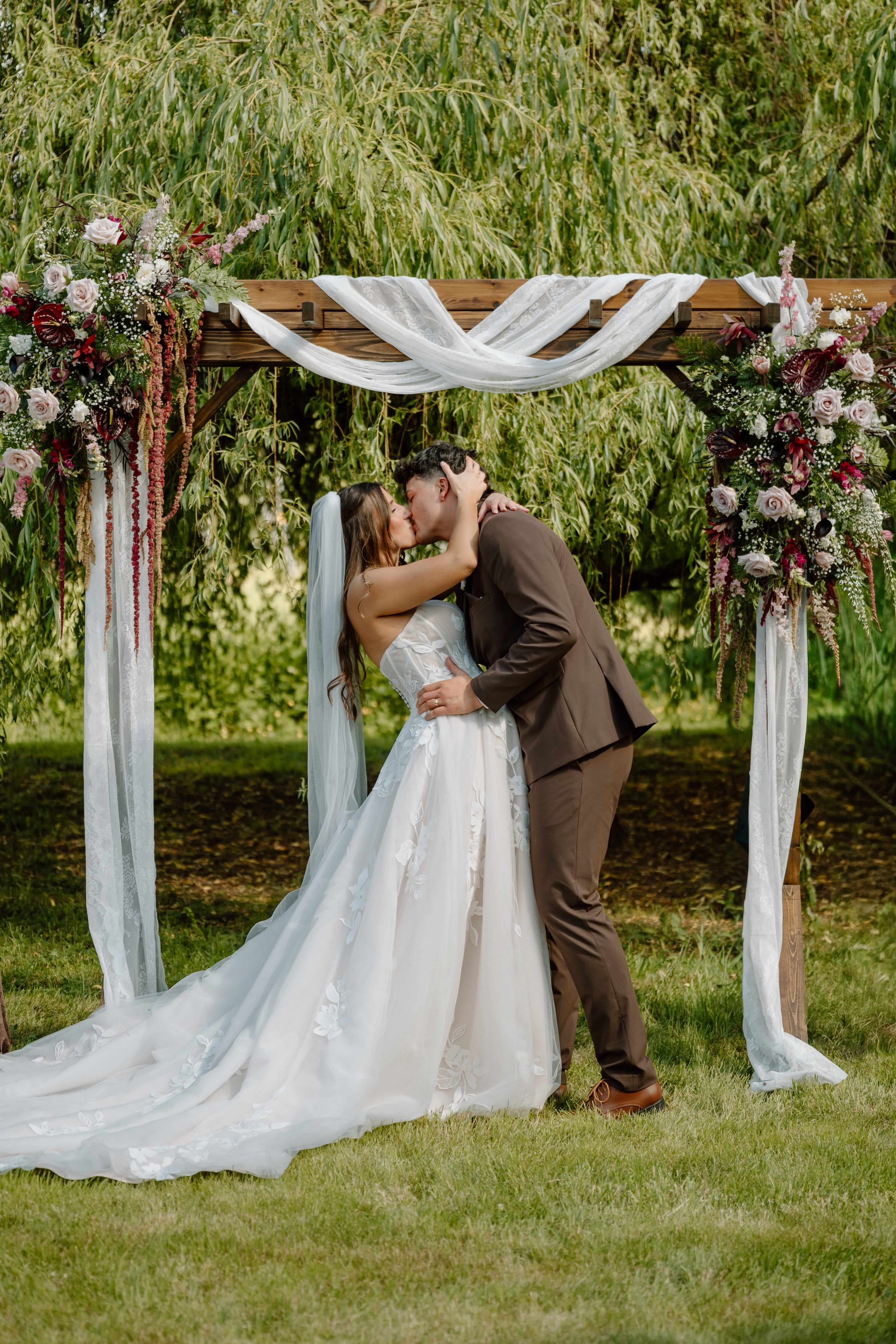 Bride and Groom at a Pacific Northwest wedding carrying lovely white and antique red cascading bouquets by Love Letter Floral and events in front of a wooden arch with beautiful draping and florals.