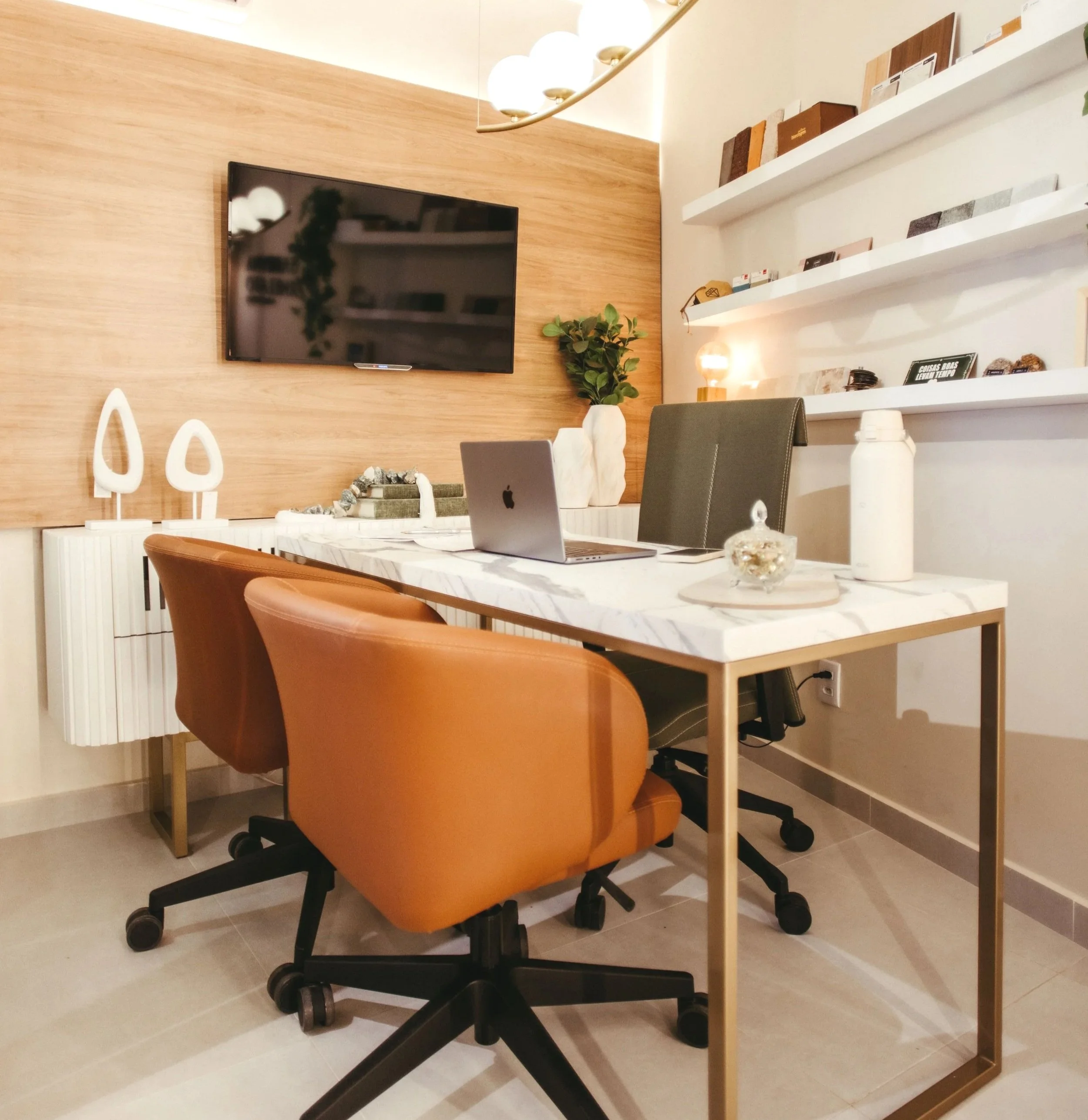 A study or office space with a white marble-top table, orange chairs, a flat-screen TV on a wooden accent wall, and white floating shelves with books and decorative items, with a white water bottle on the table. Potentially financial advisory office.