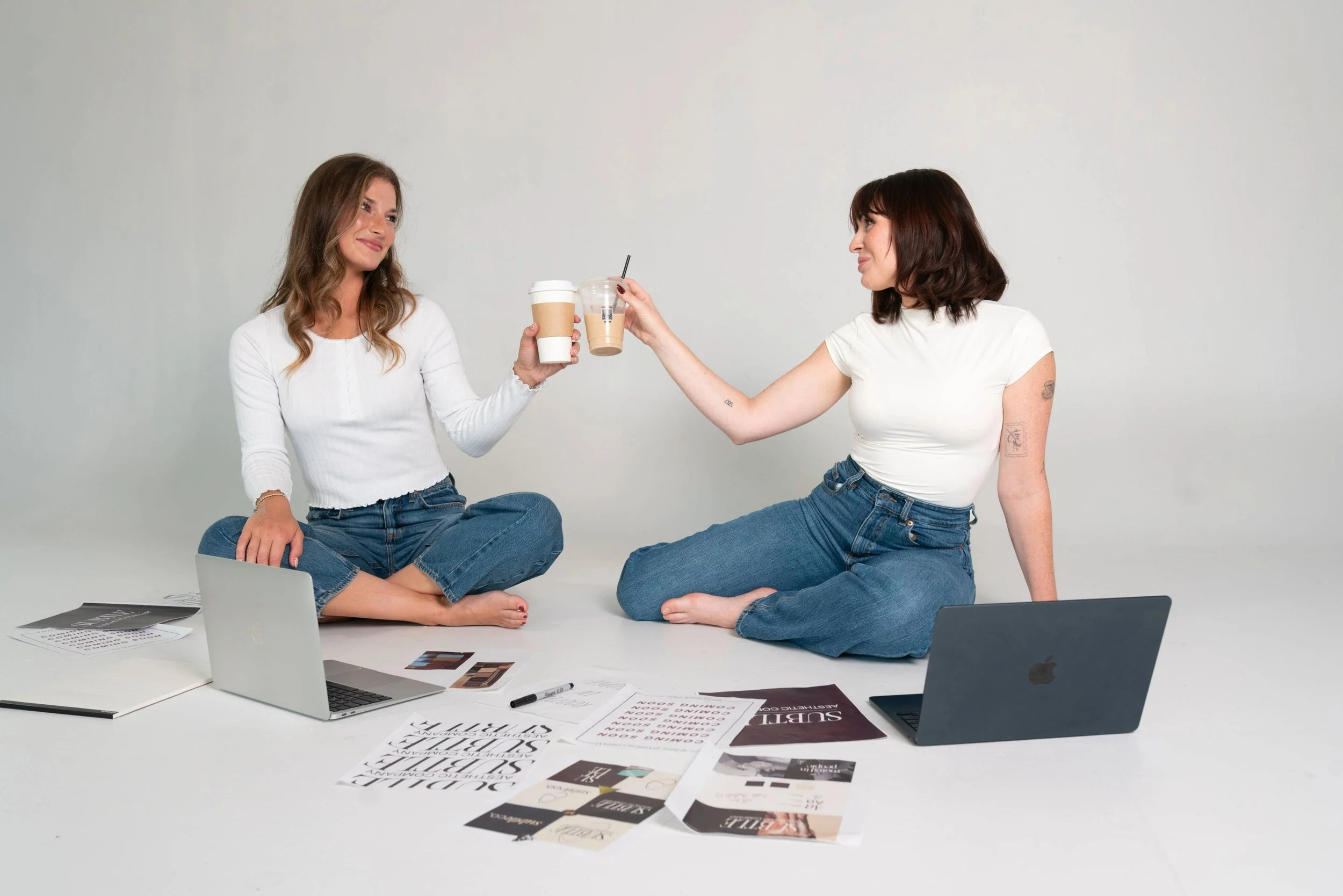 Two women sitting on the floor with laptops, papers, and coffee drinks, exchanging drinks and smiling, in a minimalistic white room.
