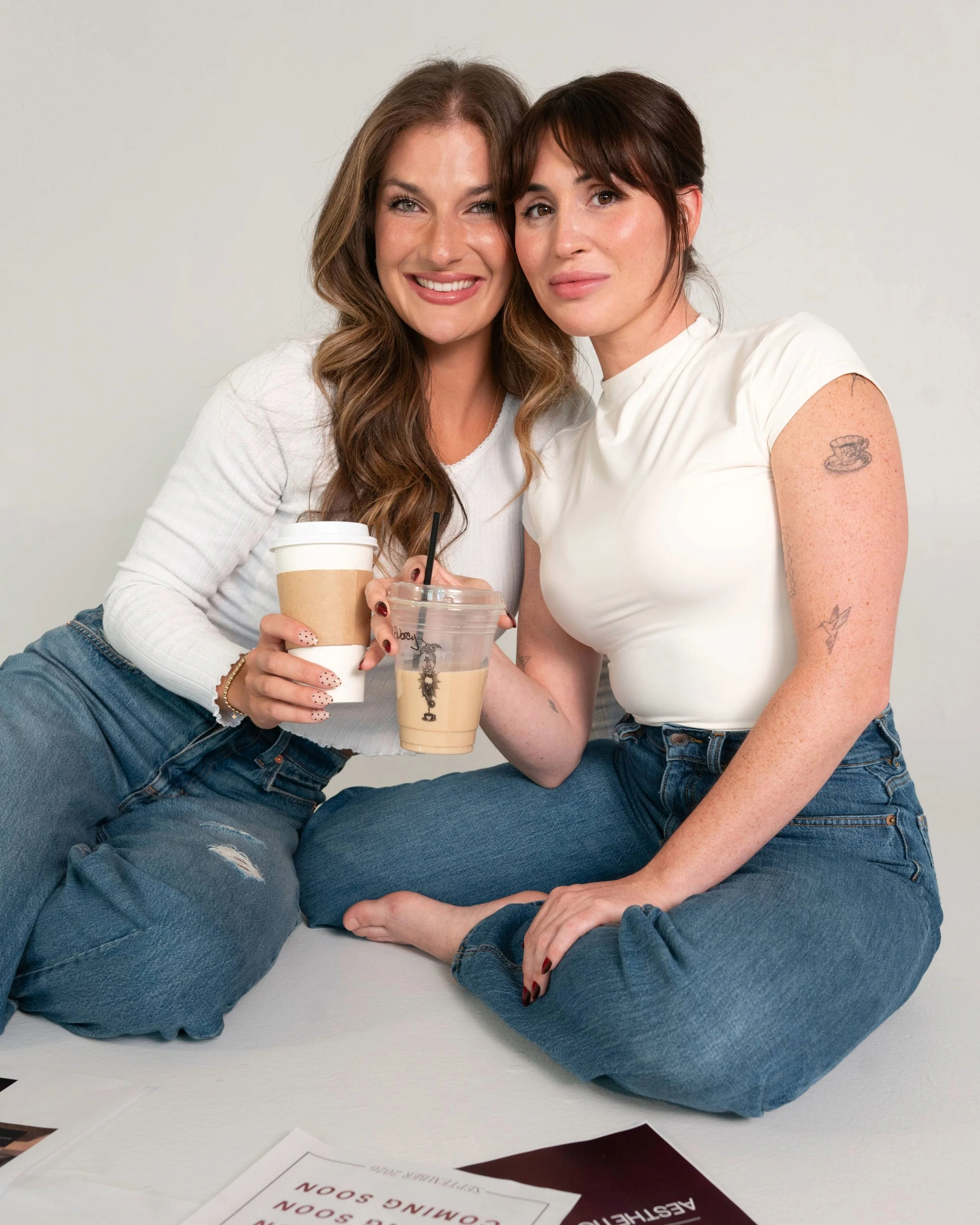 Two women sitting on the floor, holding coffee cups, smiling, and posing for the camera against a plain white background.