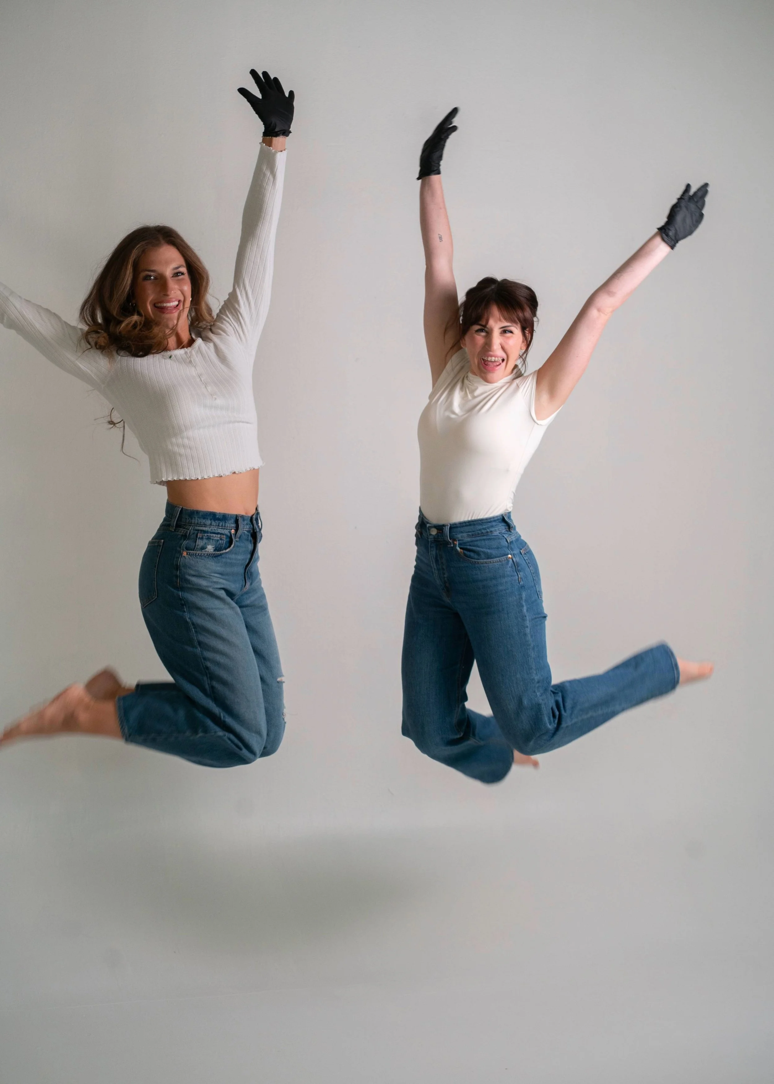 Two women jumping in the air against a plain white background, wearing casual clothes and black gloves, with happy and excited expressions.