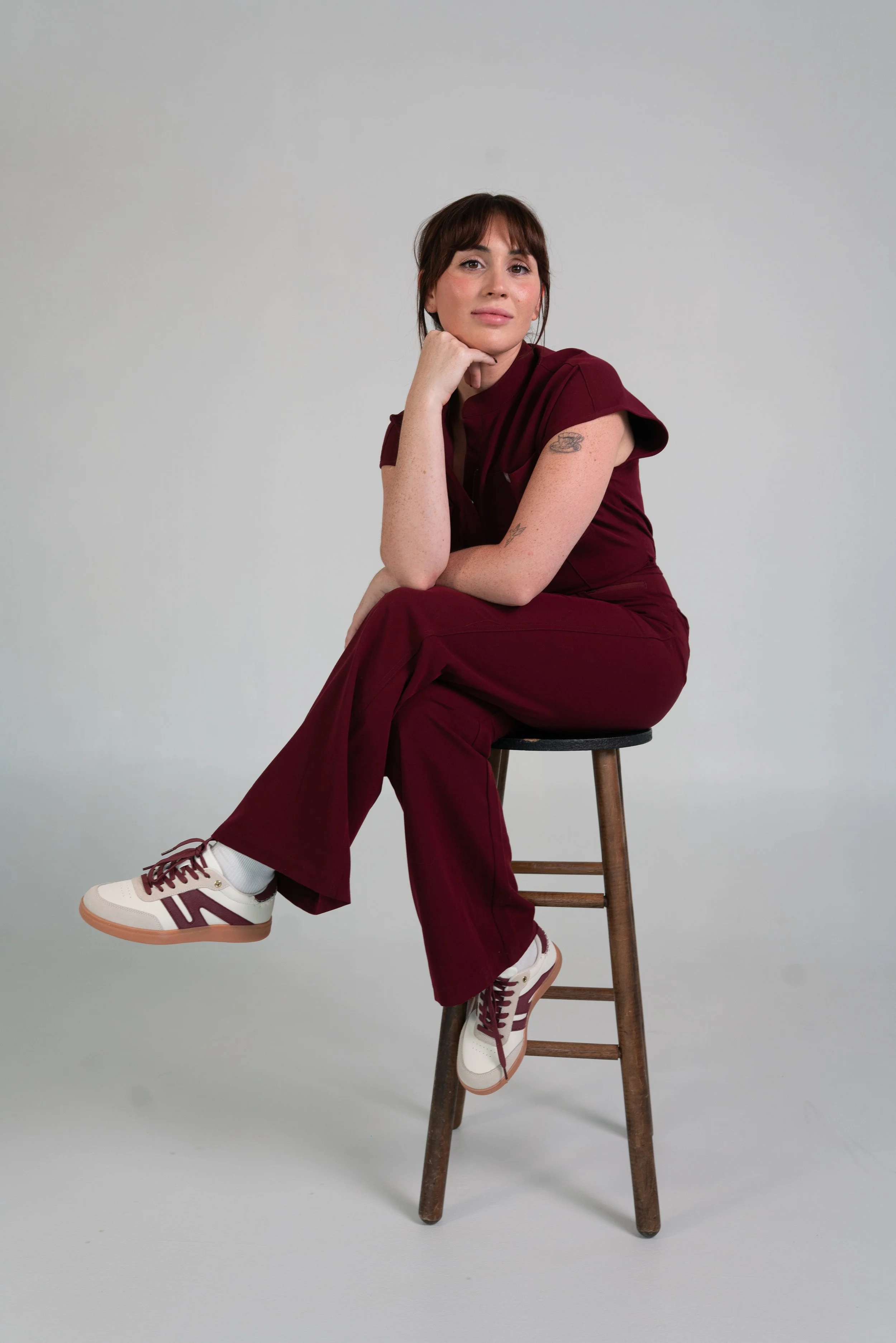 Woman sitting on a wooden stool in front of a gray background, wearing a maroon outfit and sneakers, with tattoos on her arms, resting her chin on her hand and looking at the camera.