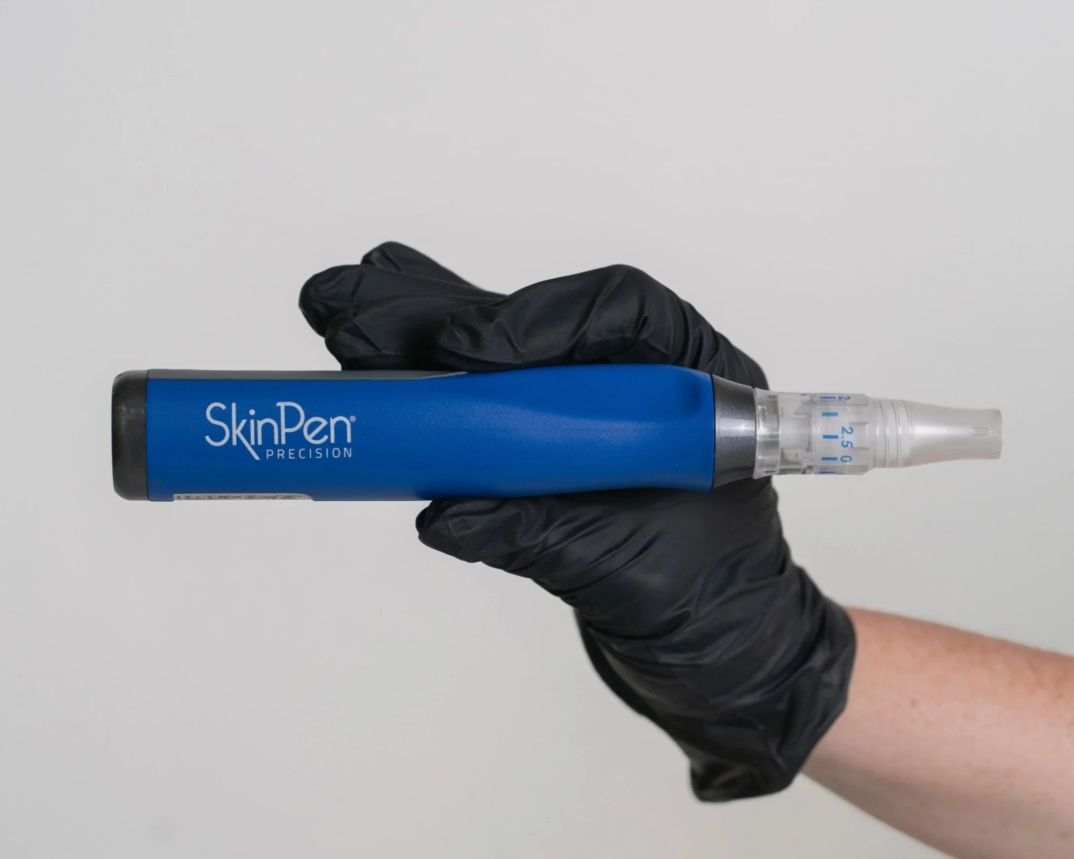 Close-up of a hand wearing a black disposable glove holding a blue SkinPen Precision device with a clear needle cartridge against a plain white background.