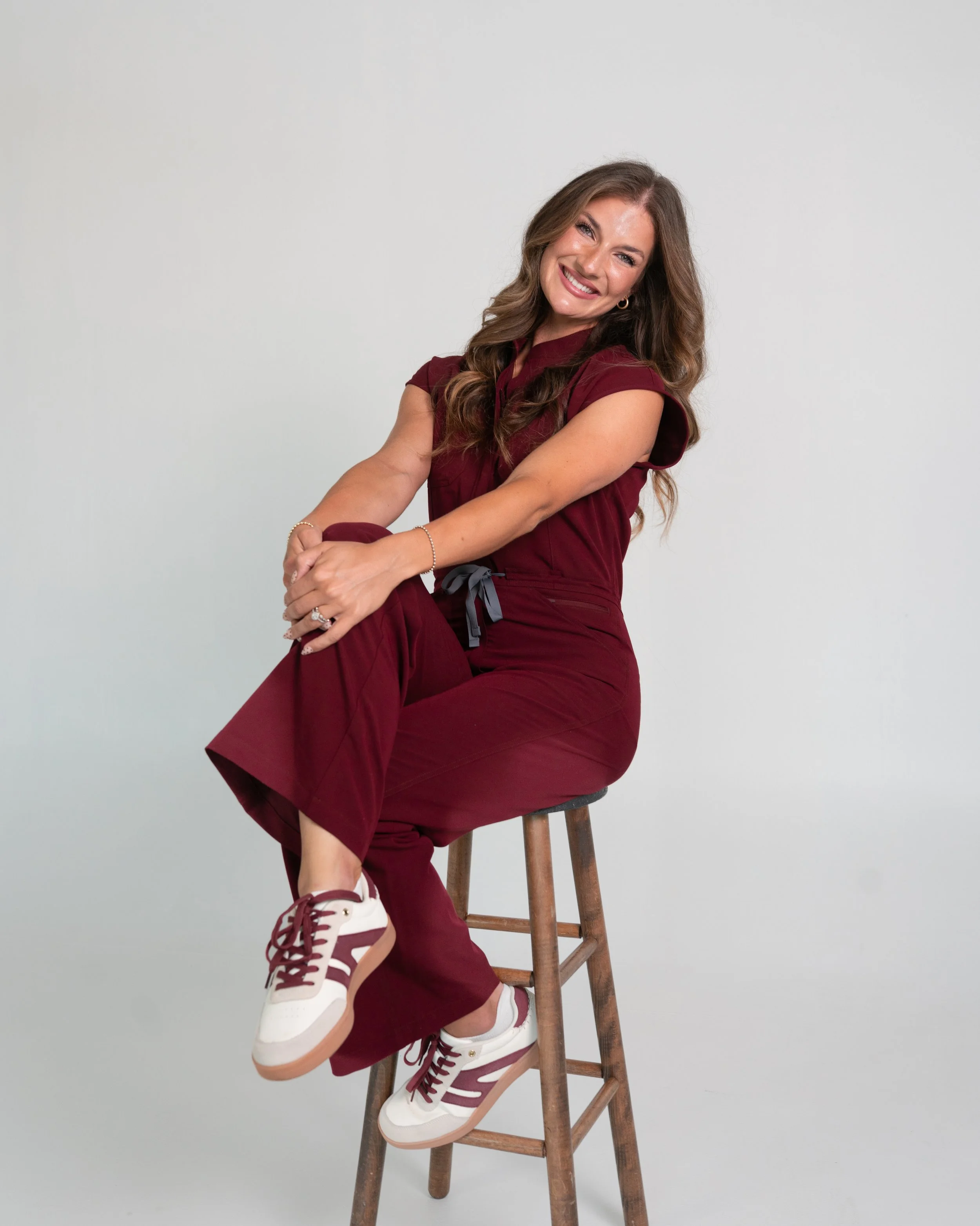 A woman with long wavy brown hair smiling, sitting on a wooden stool with one leg crossed over the other. She is wearing a burgundy outfit and white sneakers. The background is plain white.