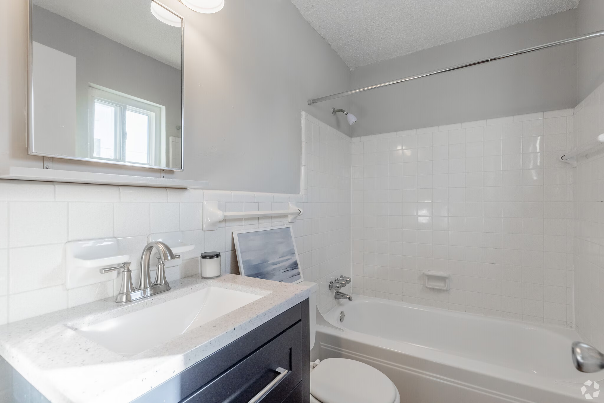 A bathroom with a white bathtub, a shower with a curtain rod, and a vanity with a sink, mirror, and gray cabinets.