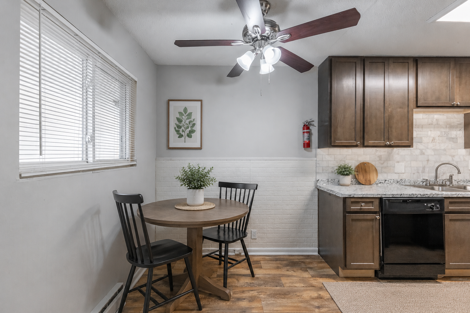 Kitchen corner with a round wooden table, two black chairs, a window with blinds, a framed leaf print on the wall, kitchen cabinets, a fire extinguisher, a potted plant, a cutting board, and a granite countertop.