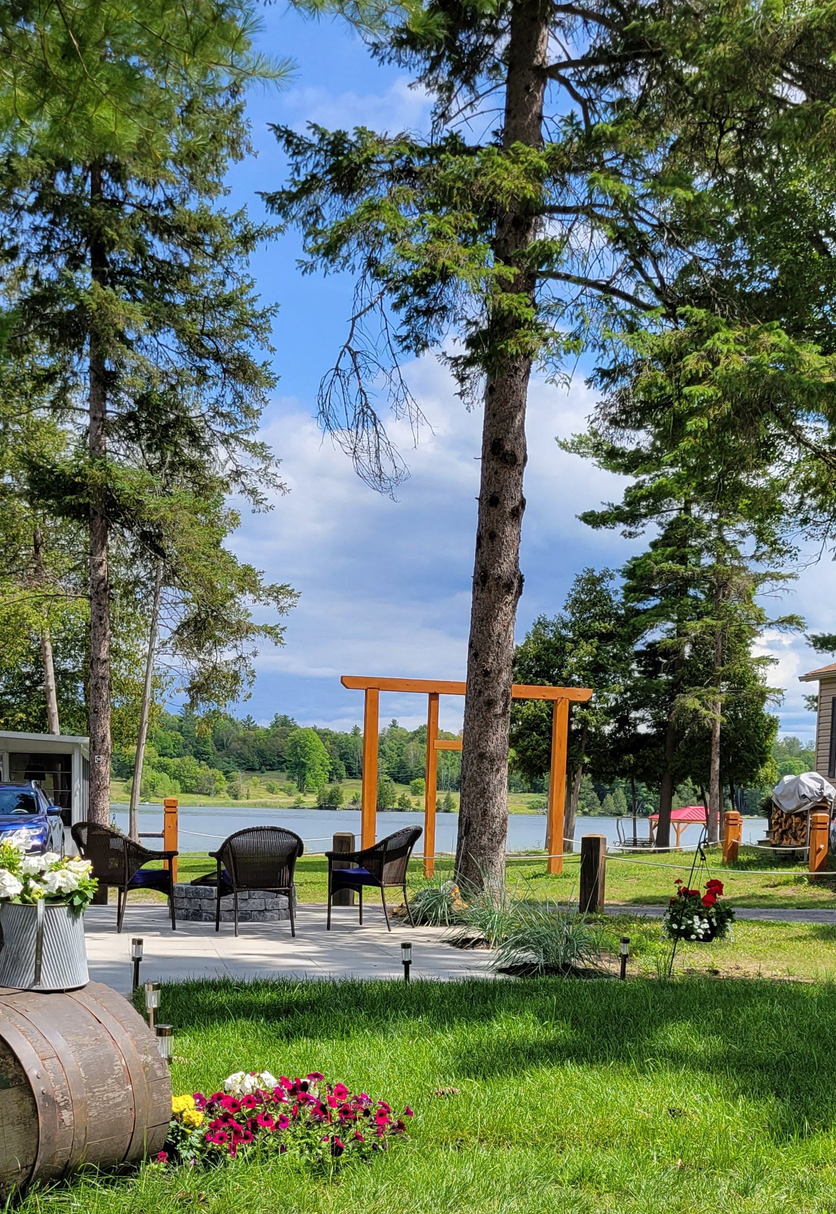 A scenic outdoor patio area near a lake with trees, flowers, outdoor chairs, and a wooden framework for a future structure.