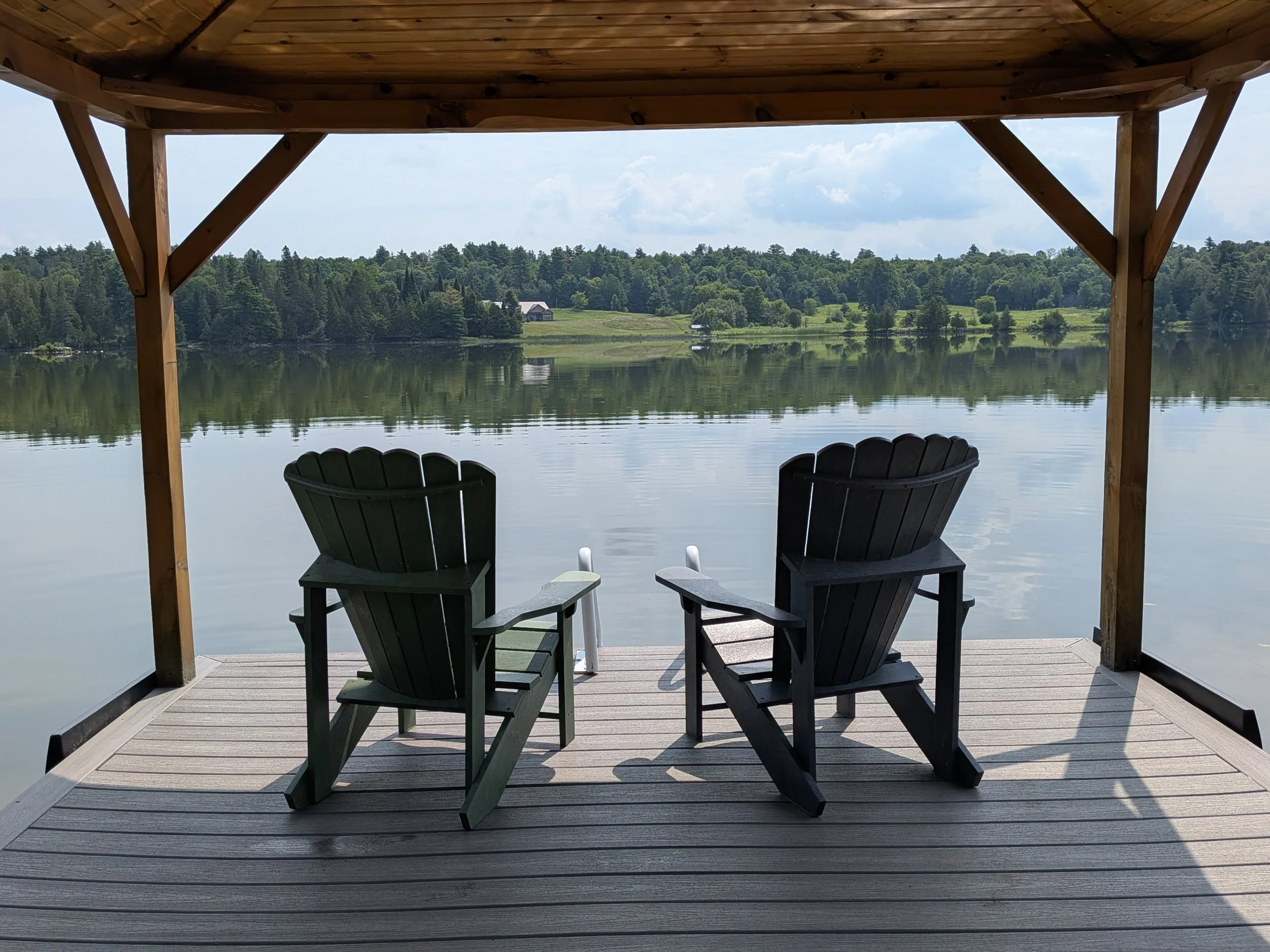 View of a lake seen from a wooden deck with two Adirondack chairs under a wooden shelter, surrounded by greenery and trees, with partly cloudy sky.
