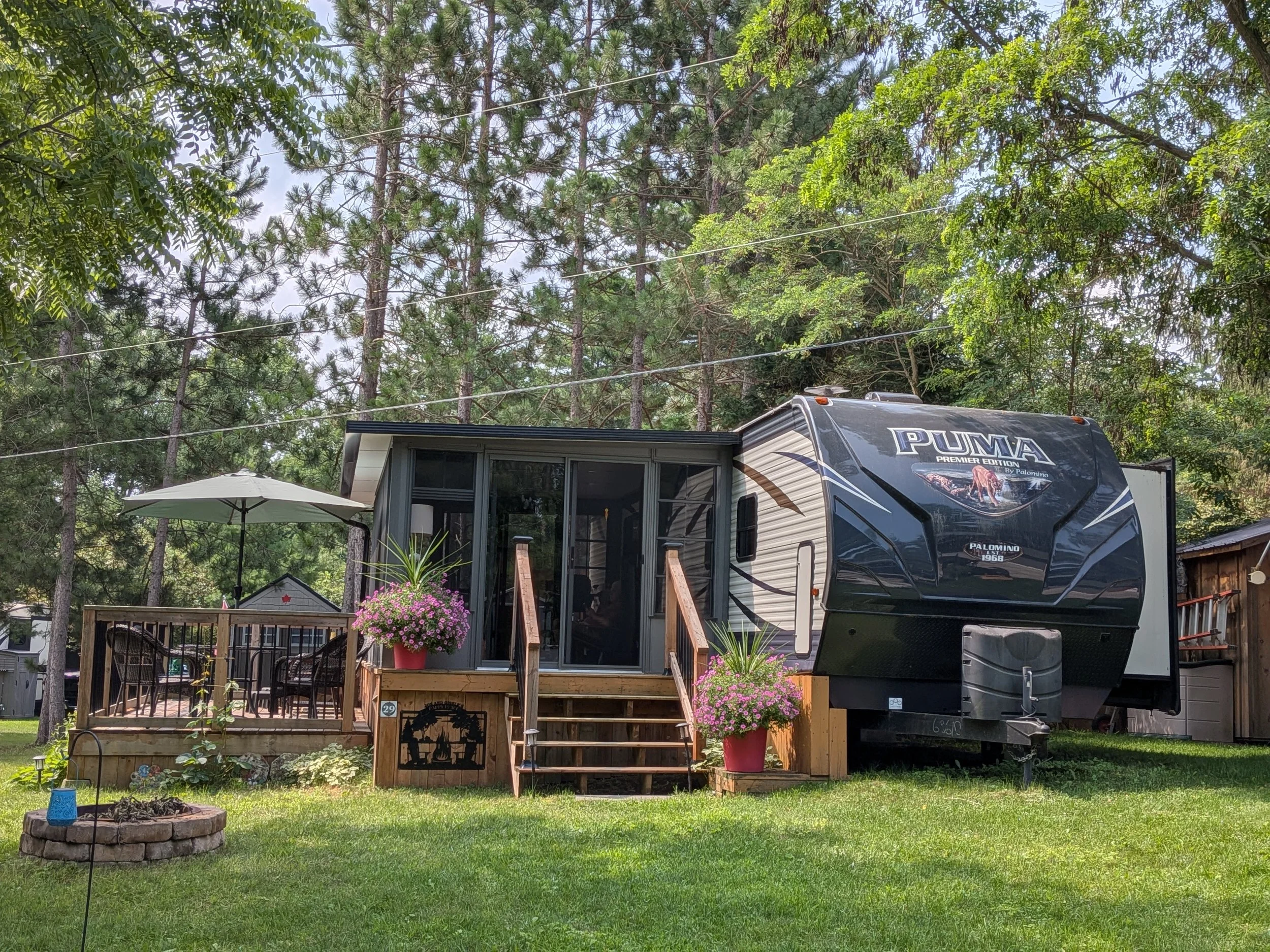 A recreational vehicle (RV) parked in a backyard with a raised deck, pink potted flowers, a white umbrella, and surrounded by tall green trees and a grassy lawn.