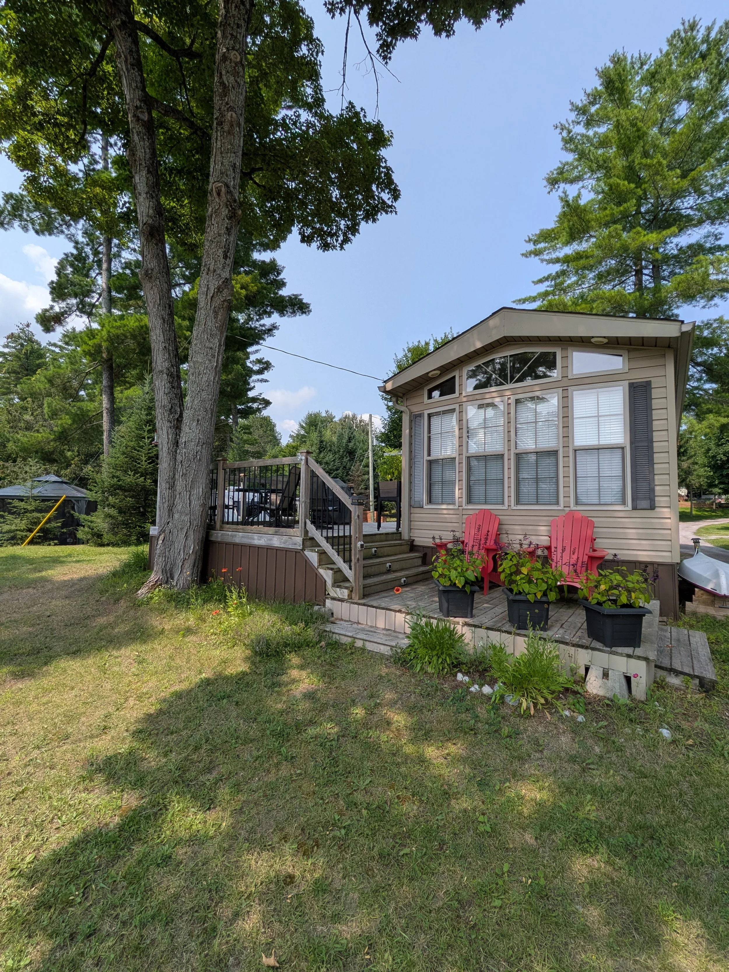 A backyard view featuring a beige house with multiple large windows, a small wooden deck with stairs, surrounded by green grass, several potted plants, tall trees including one large tree in the foreground, and a blue sky with some clouds.