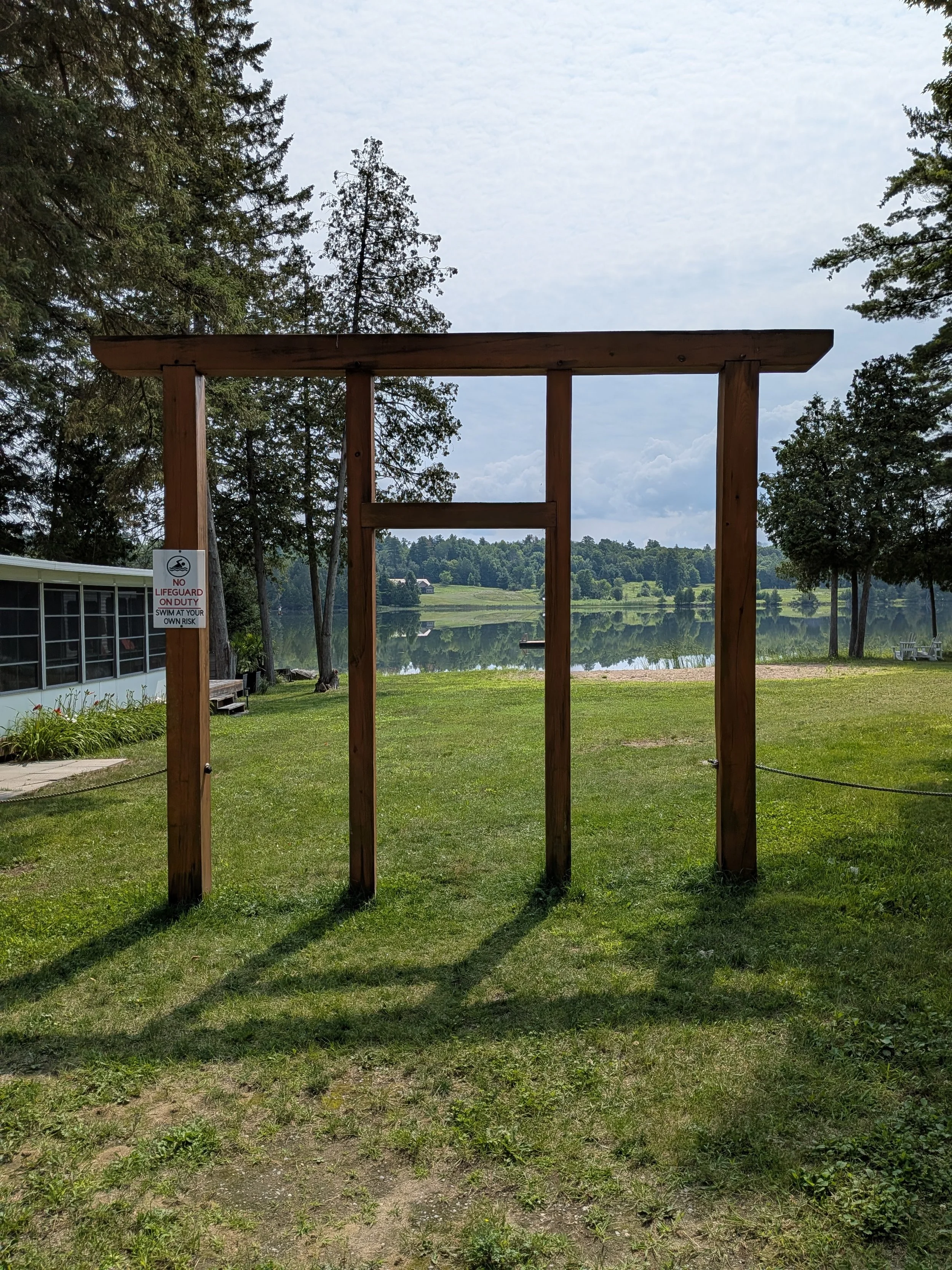 A wooden structure with a rectangular frame over a grassy area, near a lake with trees in the background, and a sign that says 'No Lifeguard On Duty. Swim At Your Own Risk.'
