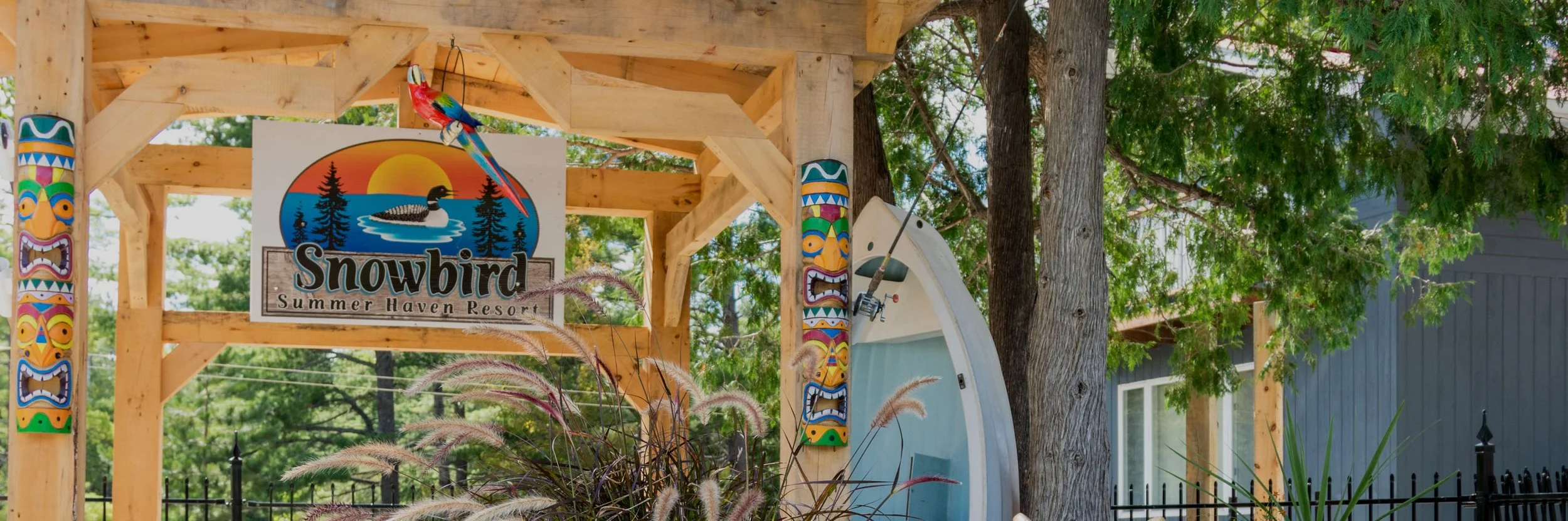 Wooden entrance sign for Snowbird Summer Haven Resort with colorful tiki-style poles, a painted sunset scene with a loon, and a real parrot perched on top, surrounded by trees and greenery.