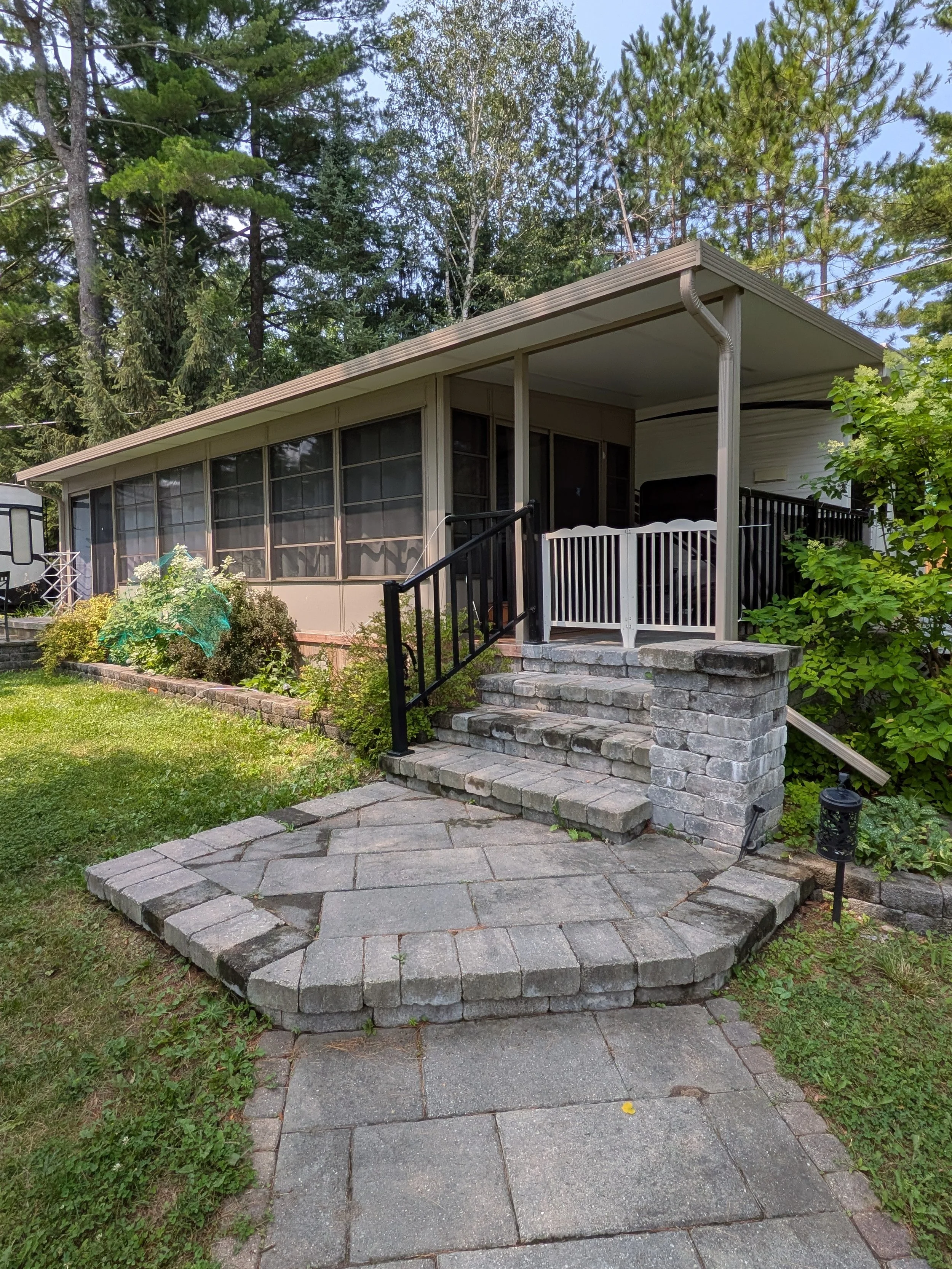 A house with a screened porch and stairs leading up to it, surrounded by a green lawn, trees, and plants, with a stone walkway in the foreground.