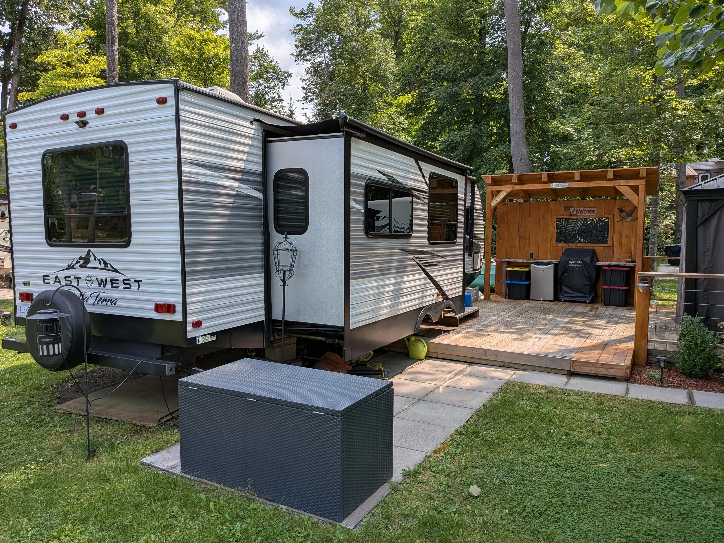 A white travel trailer with black and grey decals parked on a concrete pad in a wooded area. There is a small wooden deck attached to the trailer with a black storage bin in front of it. The background features tall trees with lush green foliage and 