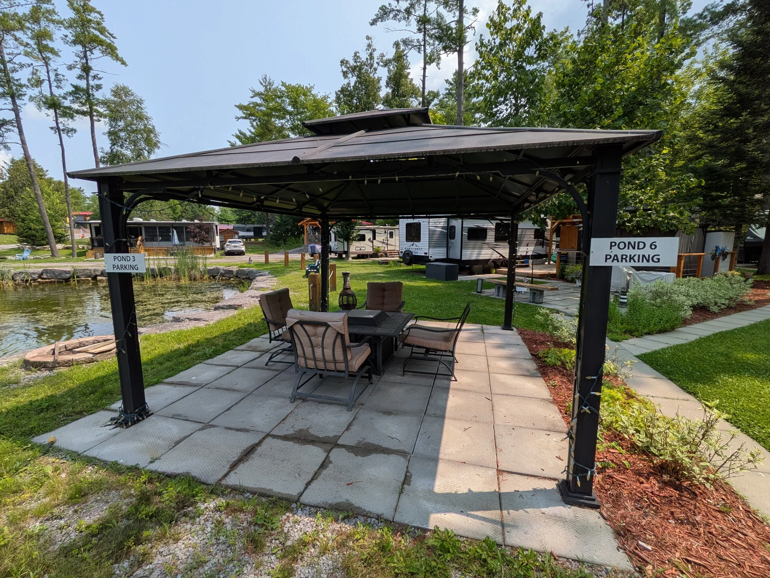 A pondside pavilion with outdoor seating, surrounded by trees, in a recreational area with RVs and scenic landscaping.