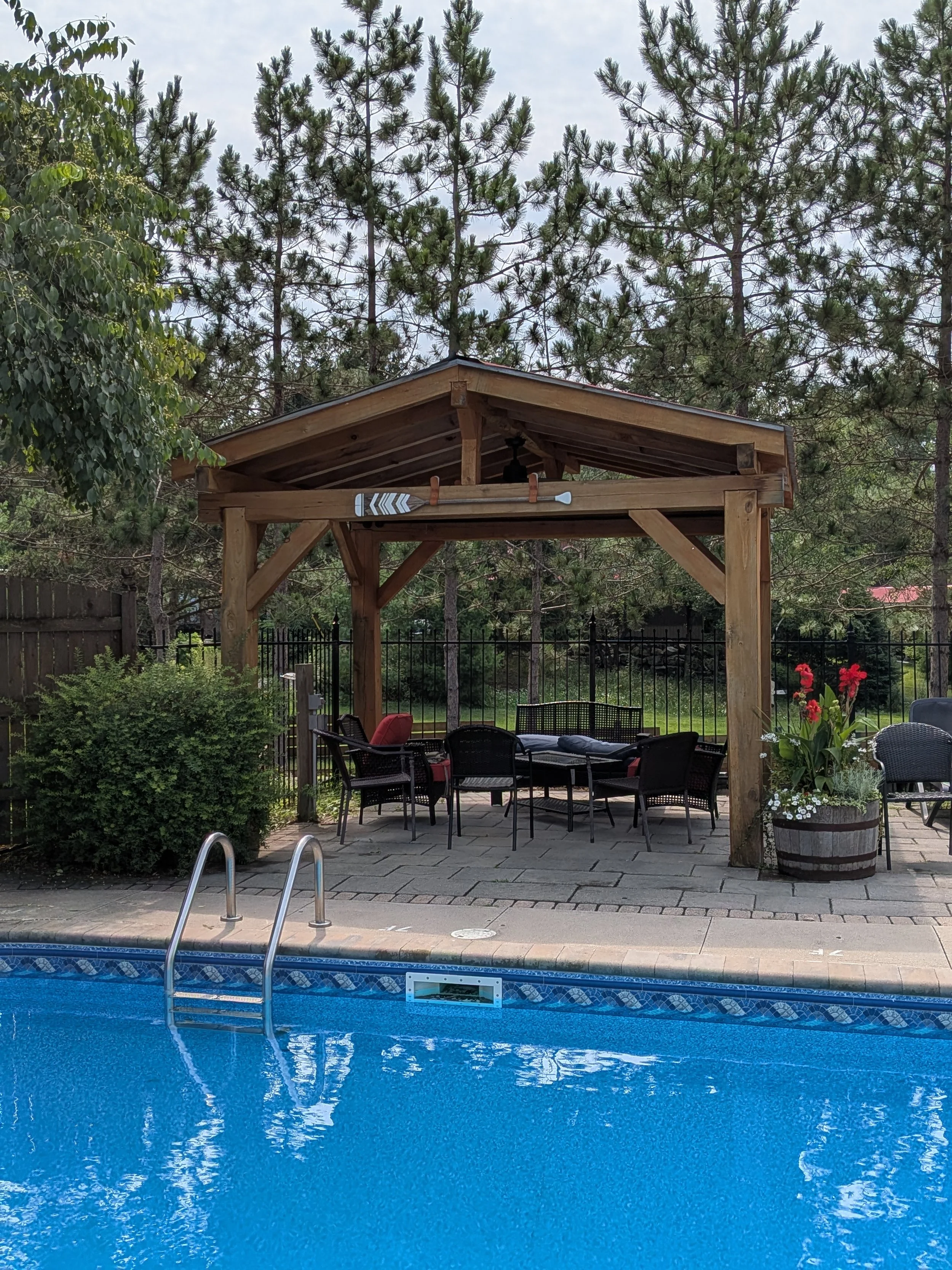 Poolside patio area with a swimming pool in the foreground, a covered seating area with chairs and a table, and a wooden pergola with trees and greenery in the background.