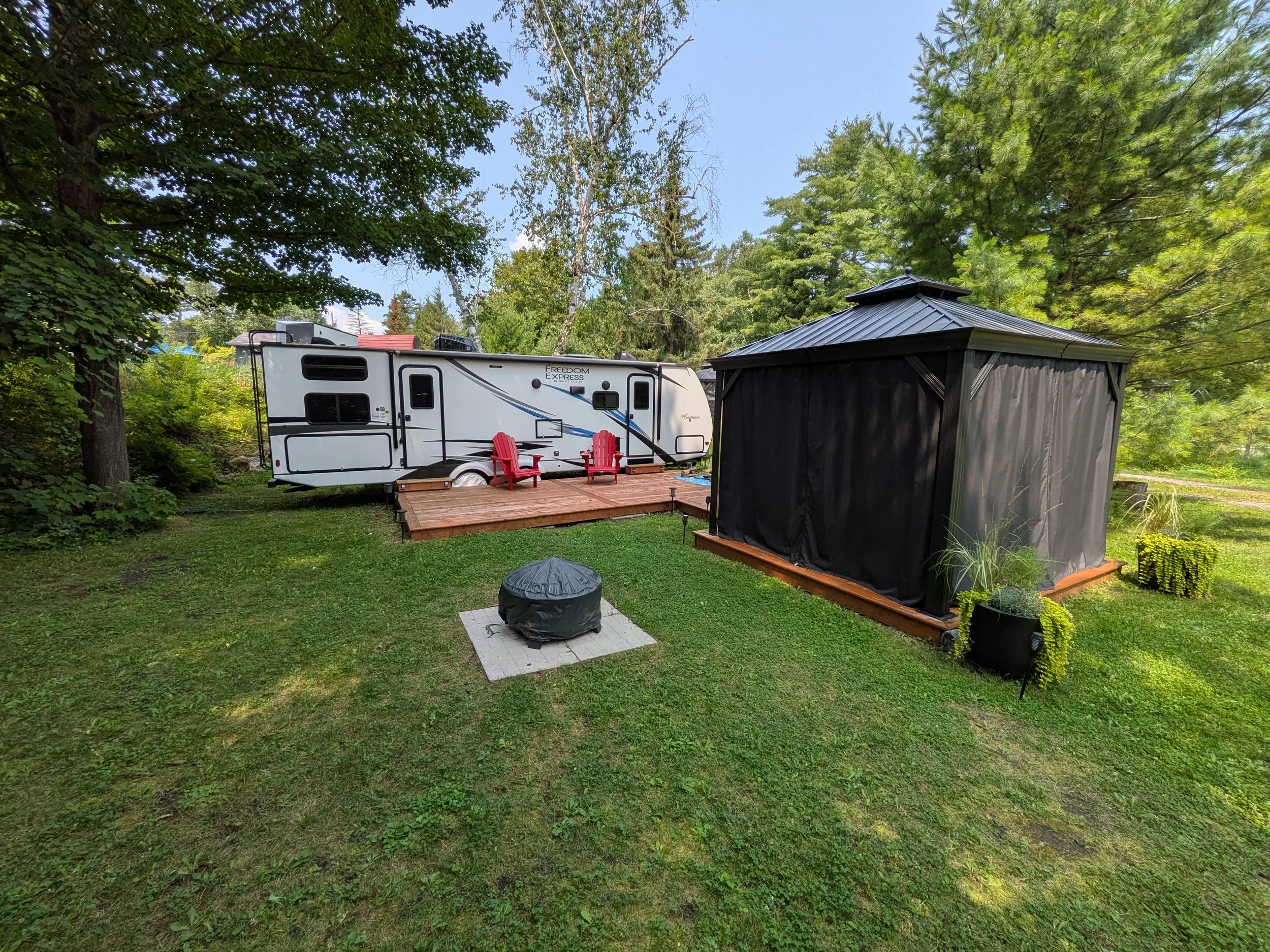 A backyard with a white travel trailer, two red chairs on a wooden deck, a black storage shed, and lush greenery with trees and grass.