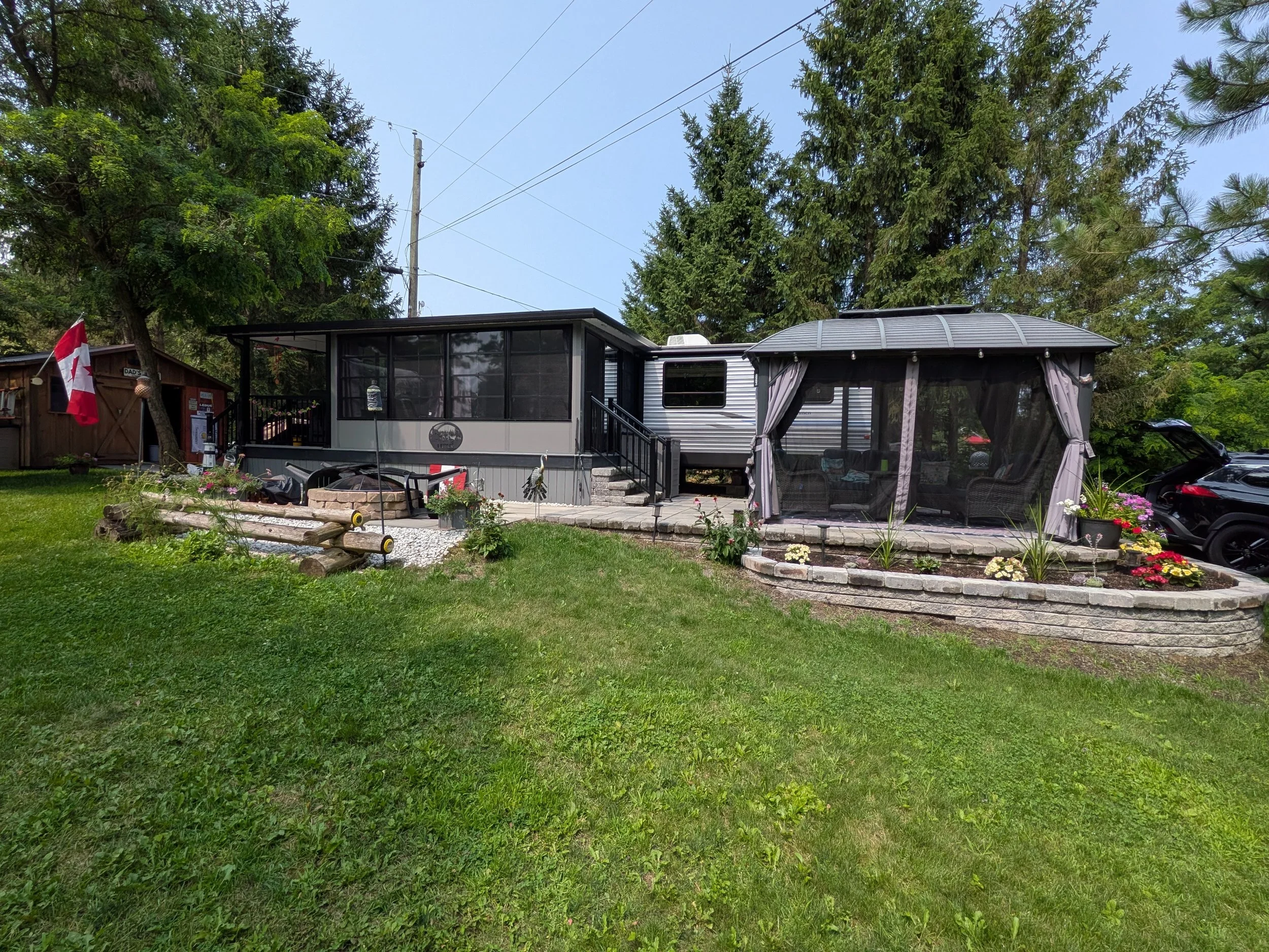 A mobile home in a green yard with trees and a small garden, with a Canada flag on a pole and a screened porch.