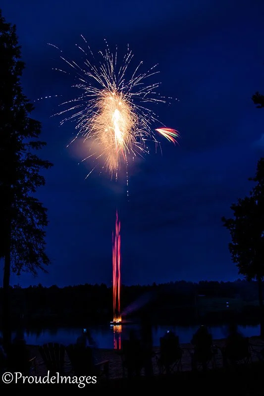 Fireworks exploding in the night sky over a body of water, with trees framing the scene and silhouettes of people watching in the foreground.