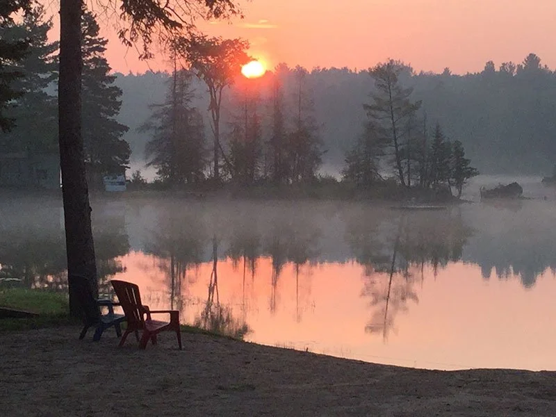 Sunrise over a calm lake with mist, surrounded by trees, with two empty chairs on the lakeshore.