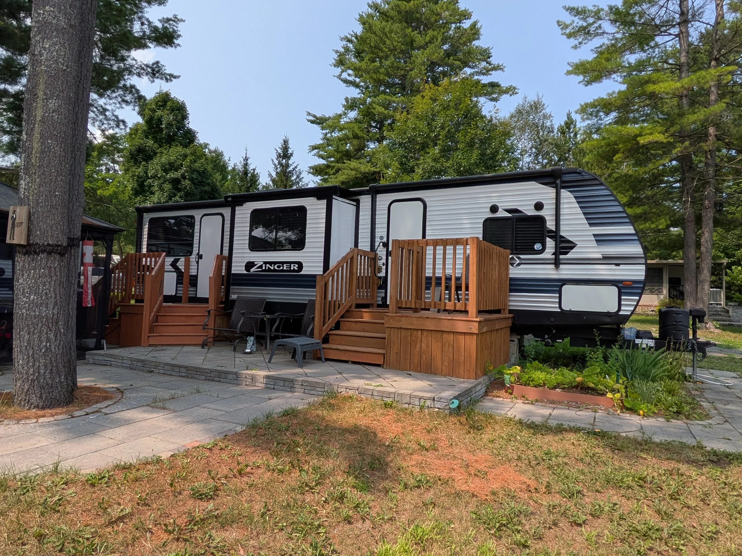 A white and black travel trailer parked among tall green trees on a clear day, with a wooden deck and outdoor seating area in front.